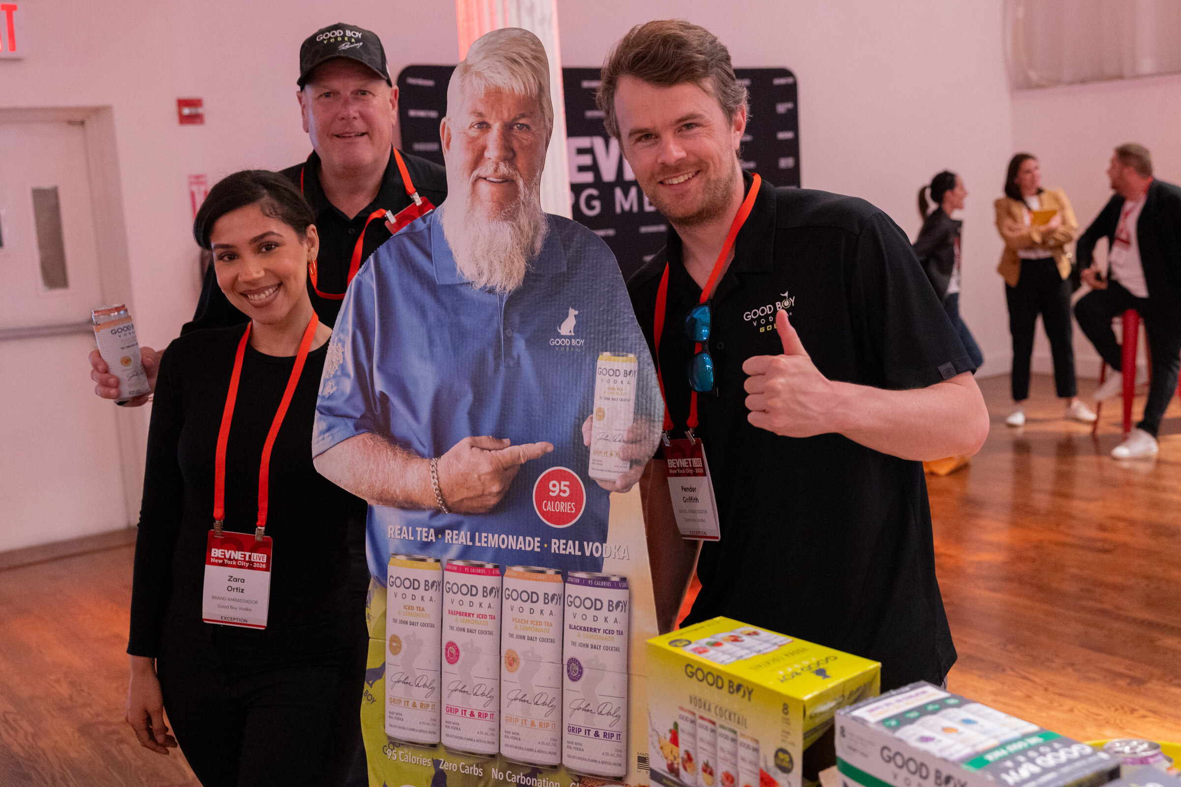 Three smiling people, one giving a thumbs-up, posing with a life-size cutout beside a Good Boy Vodka display.