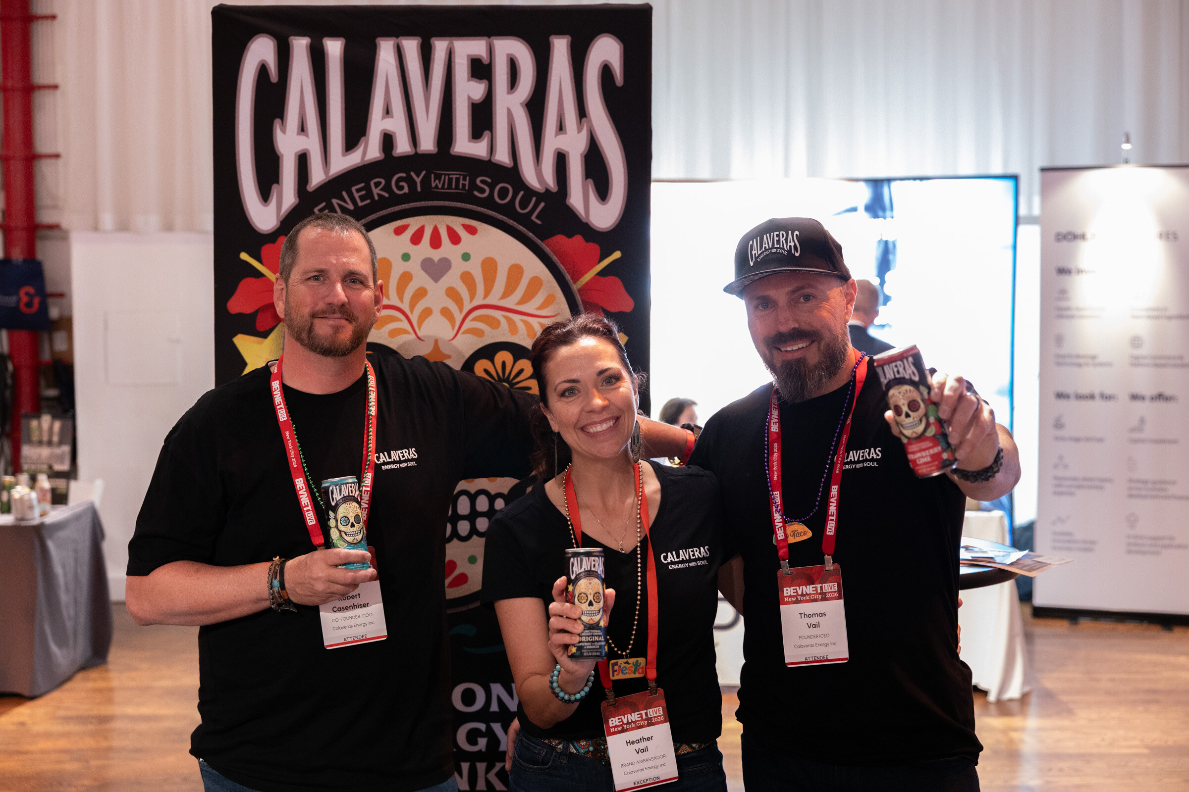Three people in black Calaveras shirts holding up cans in front of a Calaveras Energy sugar skull banner.