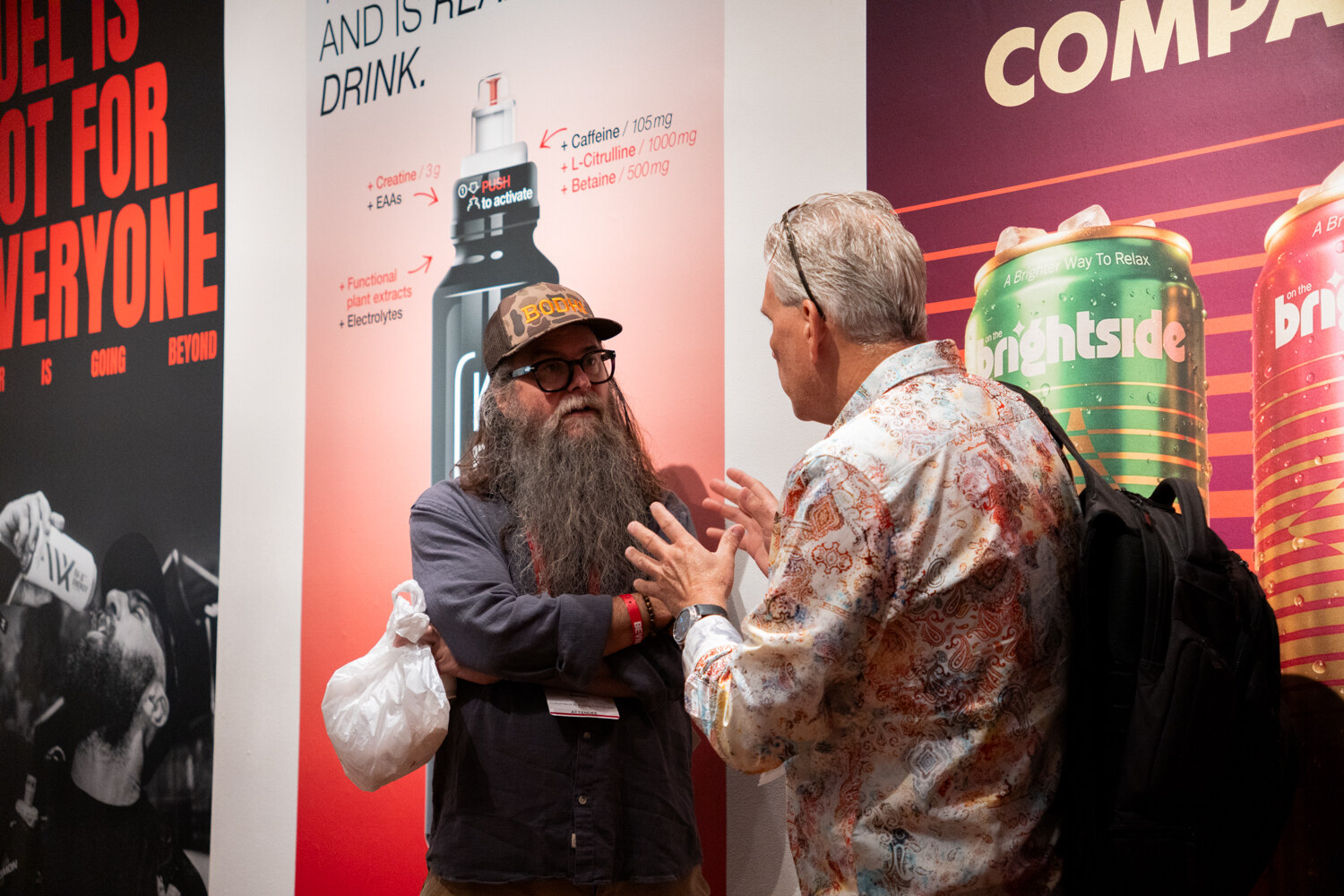 Bearded man in a cap talking with a man in a patterned shirt in front of colorful sponsor posters including a Brightside can.