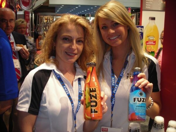 Two smiling women in white and black Fuze polo shirts and lanyards hold bottles of Fuze beverages. A Minute Maid orange juice bottle is visible on a display behind them.