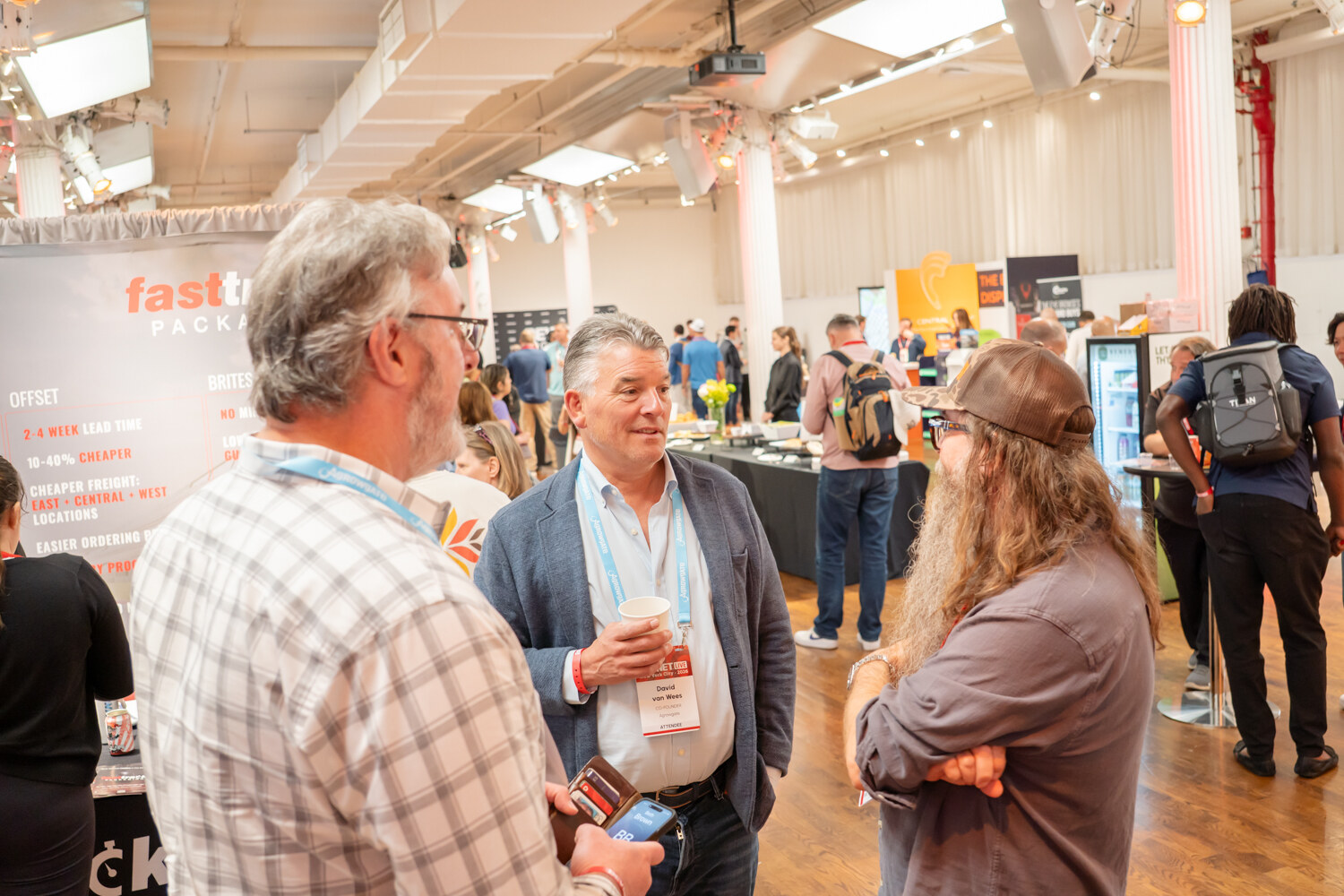Group of attendees, one holding a coffee cup, talking on the expo floor with a fasttrack Packaging sign behind.