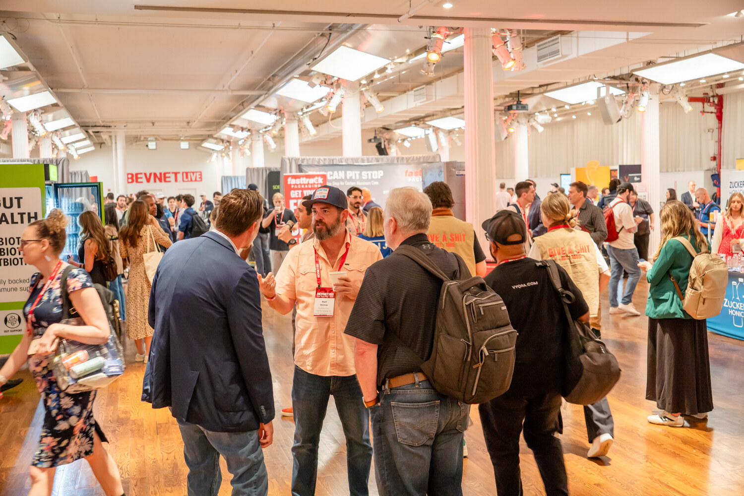 Attendees walking and talking among sponsor booths in a bright hall with a BevNET Live sign and a Can Pit Stop banner in the background.