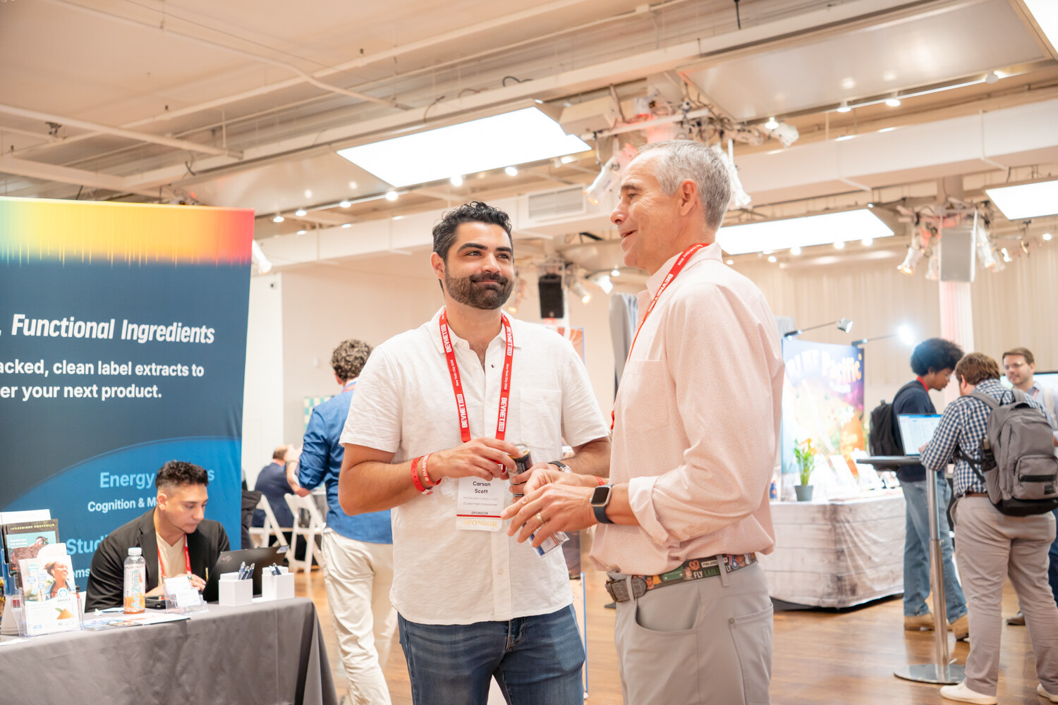 Two smiling men with lanyards talking next to a booth banner for functional ingredients.