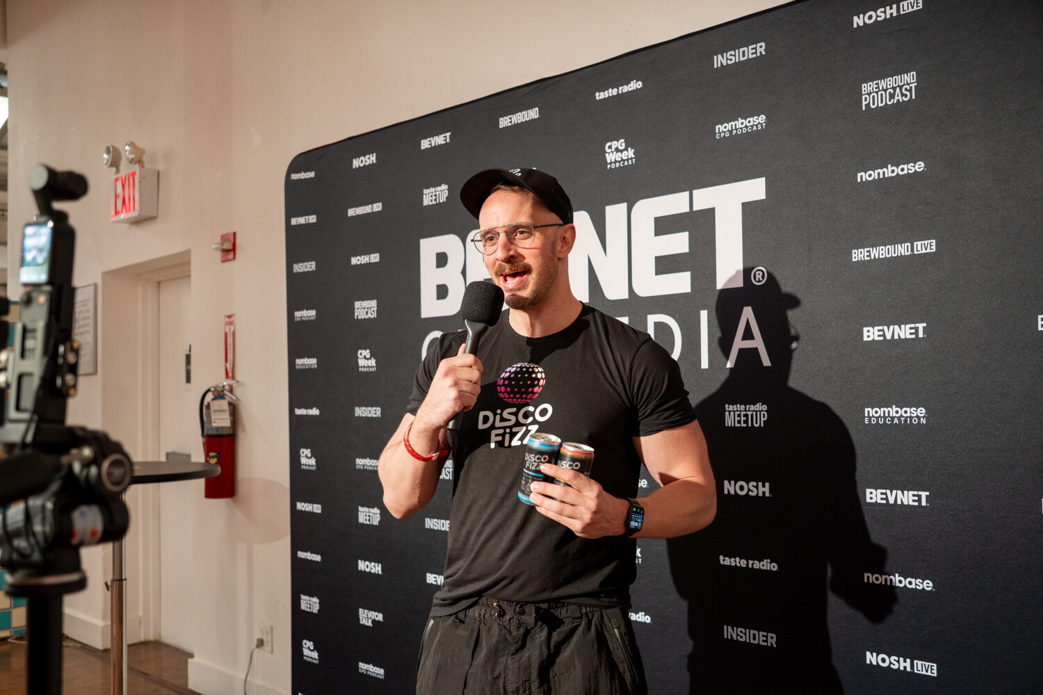 Man in a Disco Fizz T-shirt and cap speaking into a microphone while holding two Disco Fizz cans in front of a BevNET CPG Media backdrop.
