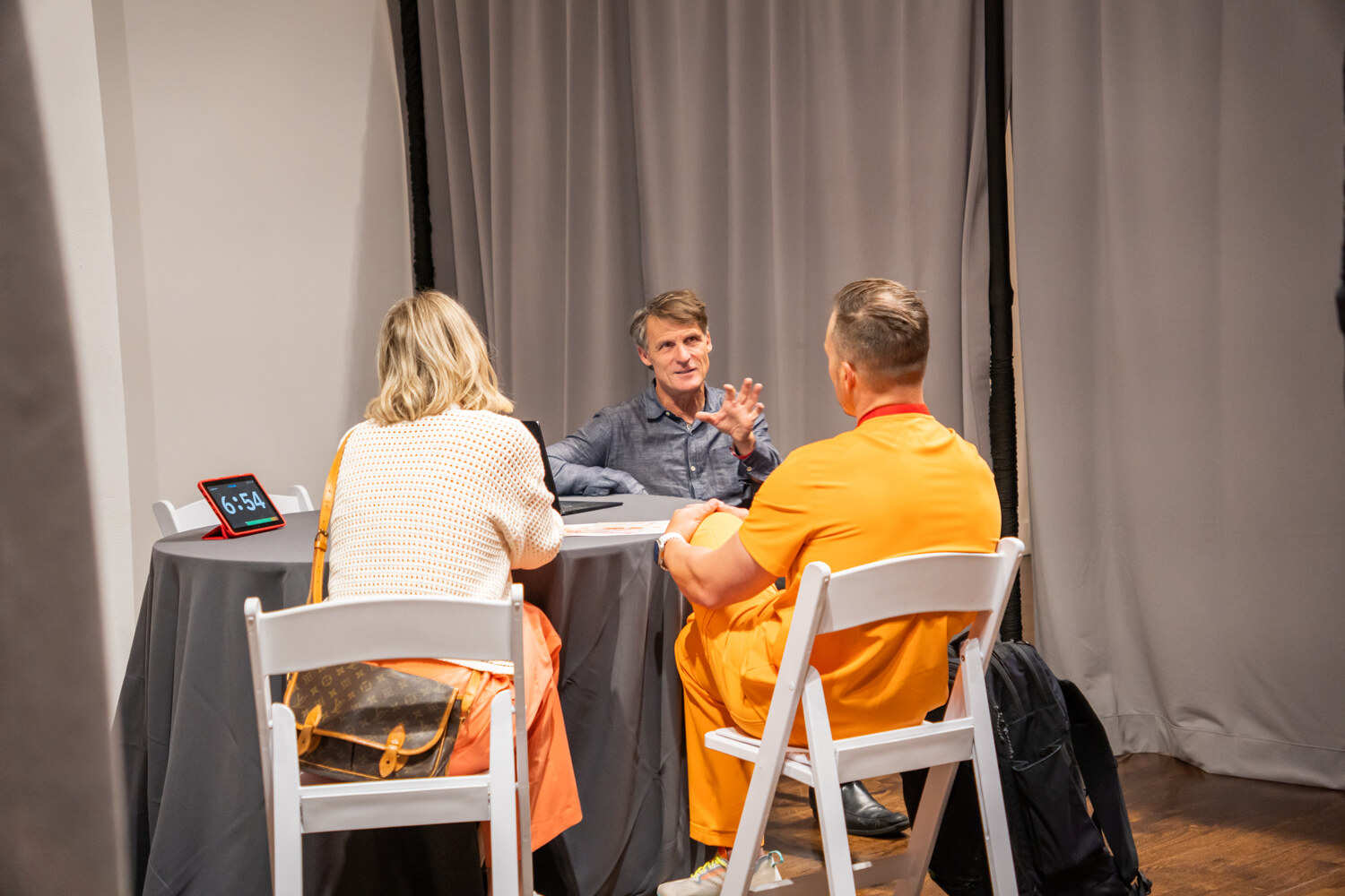 Man in a gray shirt gesturing toward two attendees, one in an orange outfit, at a round table with a timer.