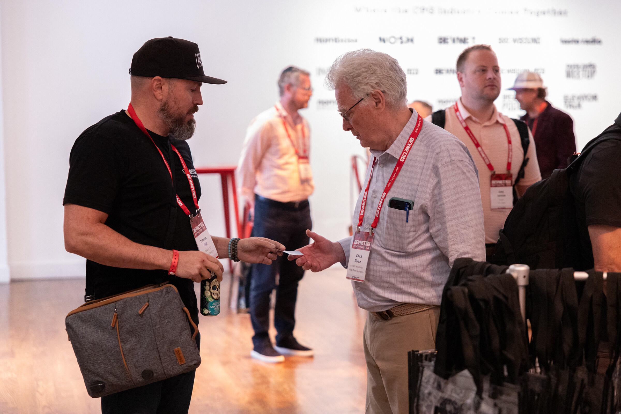 Man in a black cap handing a business card to an older attendee in a checked shirt.