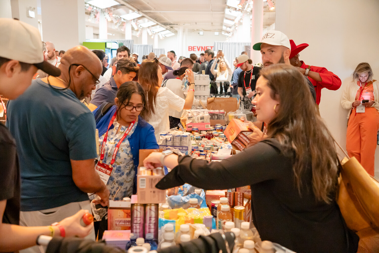 Attendees reaching across a long table crowded with beverage cans, bottles and boxes of samples.