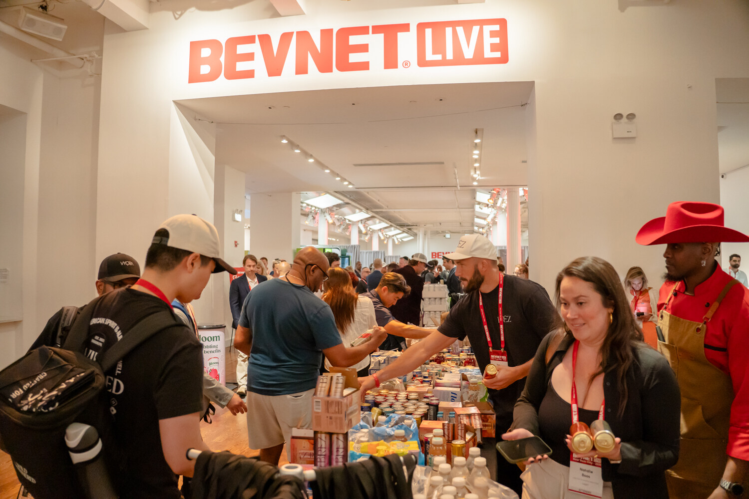 Attendees gathering samples from a long table of drinks under a BevNET Live sign, one woman smiling as she holds two bottles.