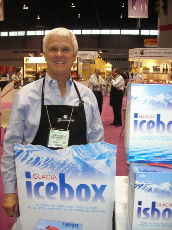 A smiling white-haired man in a black apron and blue shirt stands behind boxes of Glacia Icebox Arctic spring water. European Imports booths and other attendees are in the hall behind him.
