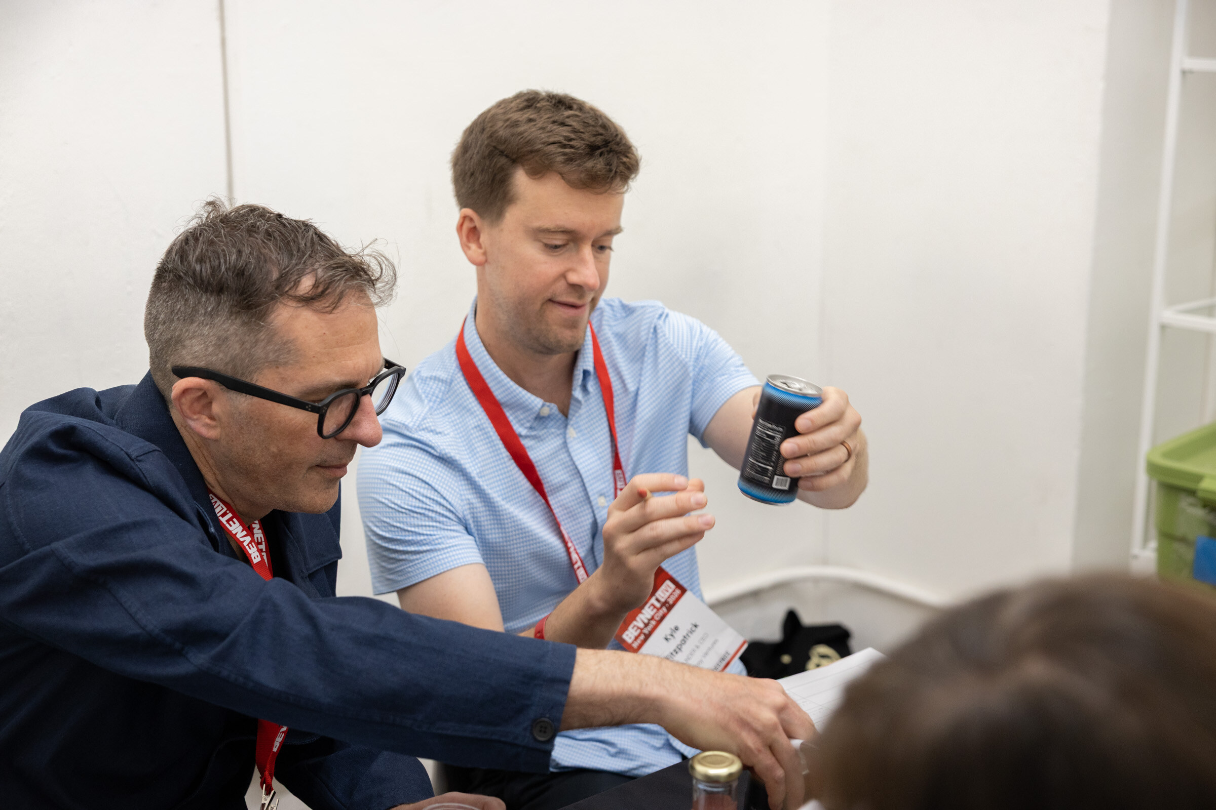 Man in glasses and a man in a light blue shirt with lanyards pour from a can into sample cups at a judging table.