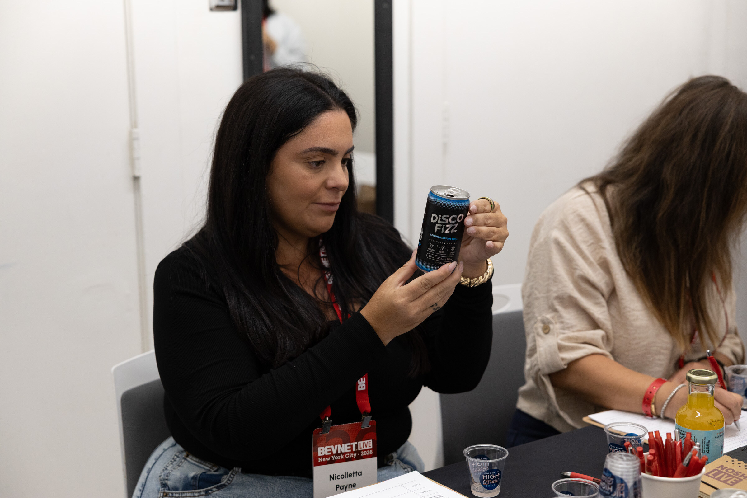 Woman with a BevNET Live lanyard holds a dark can of Disco Fizz at a judging table beside another judge.