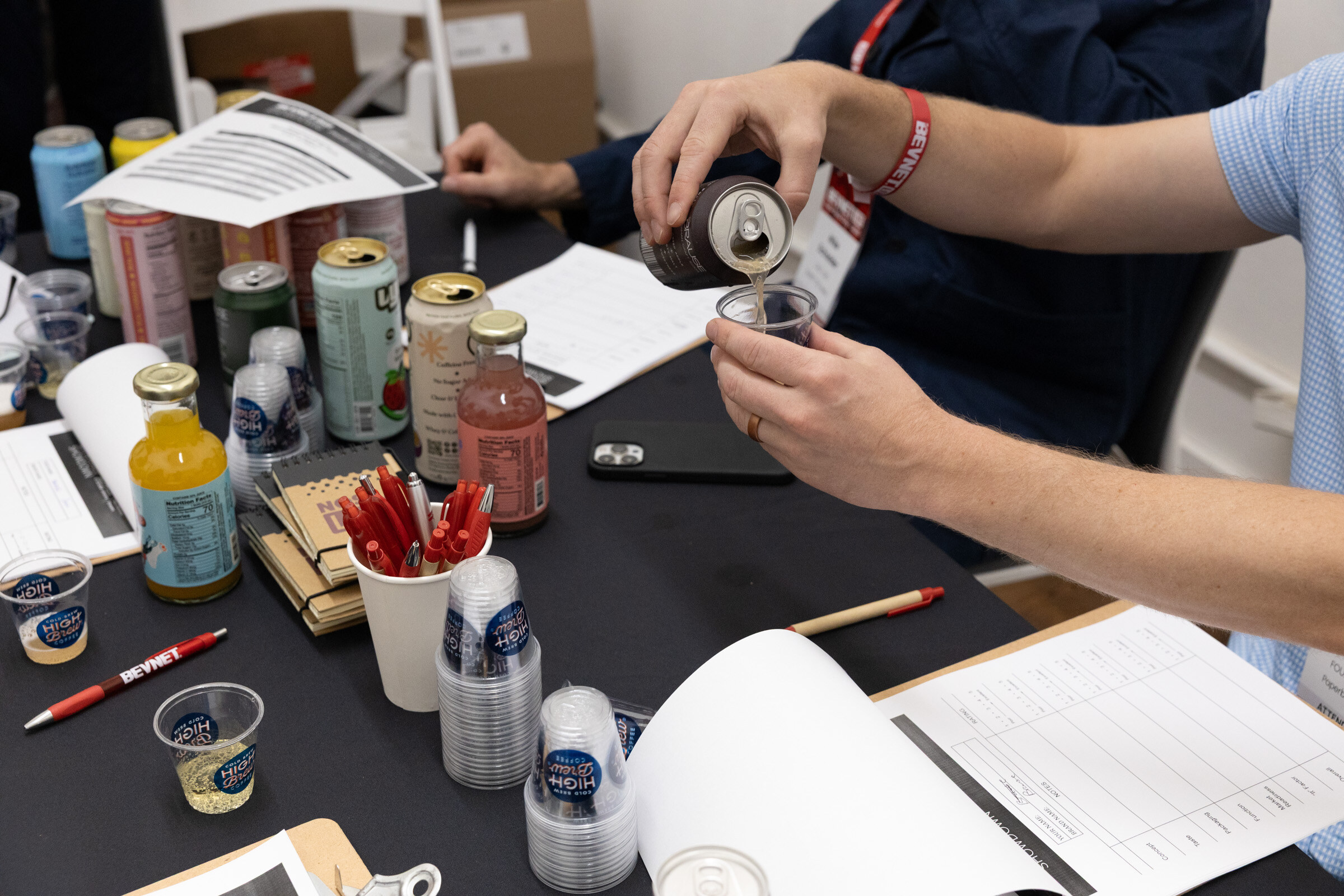 Close-up of hands pouring a can into a plastic sample cup over a black table with cans, bottles, pens and score sheets.