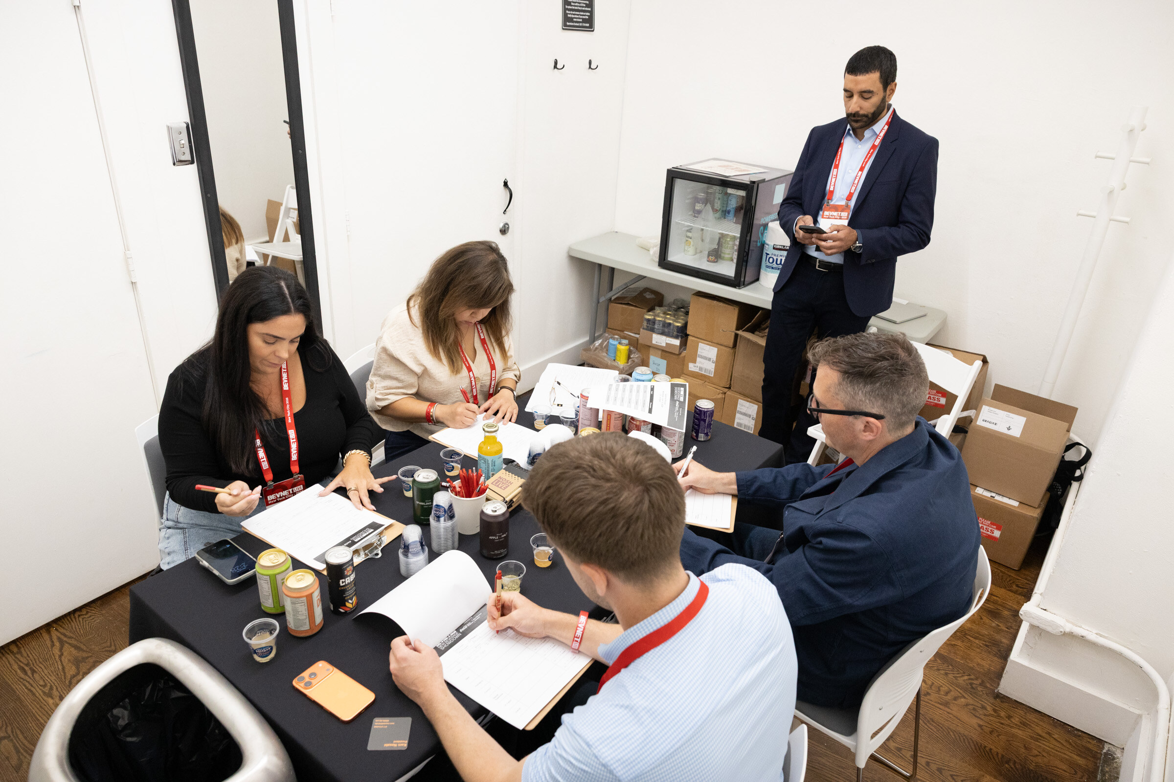 Four judges with lanyards sit around a table of cans and score sheets while a host in a suit stands nearby in a small white room.