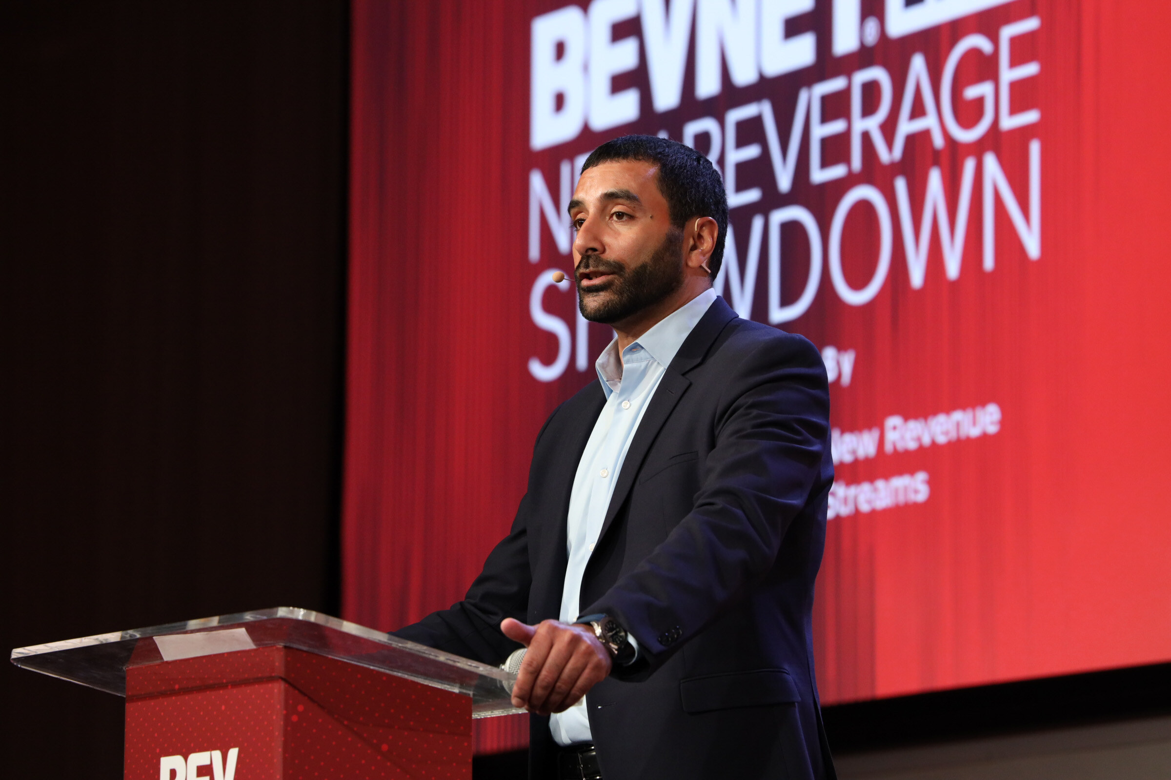 Man in a navy suit speaks at a BevNET Live podium in front of a red New Beverage Showdown screen.