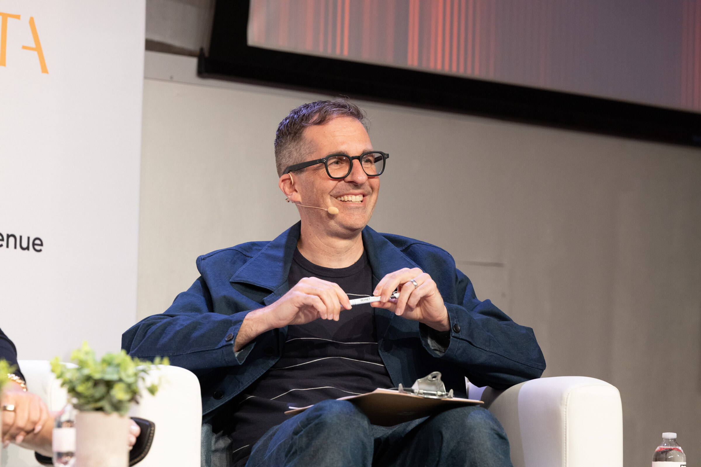 Man in black glasses and a navy jacket smiles from a white couch, holding a pen over a clipboard.