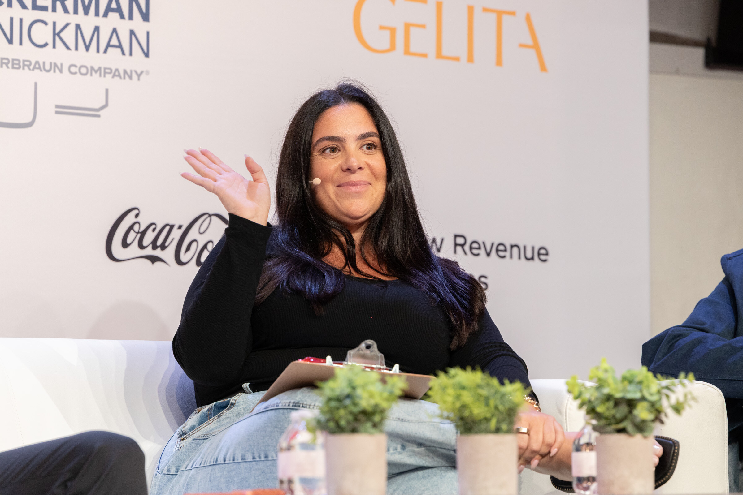 Smiling woman with long dark hair waves from a white couch, a clipboard on her lap, in front of a sponsor wall.