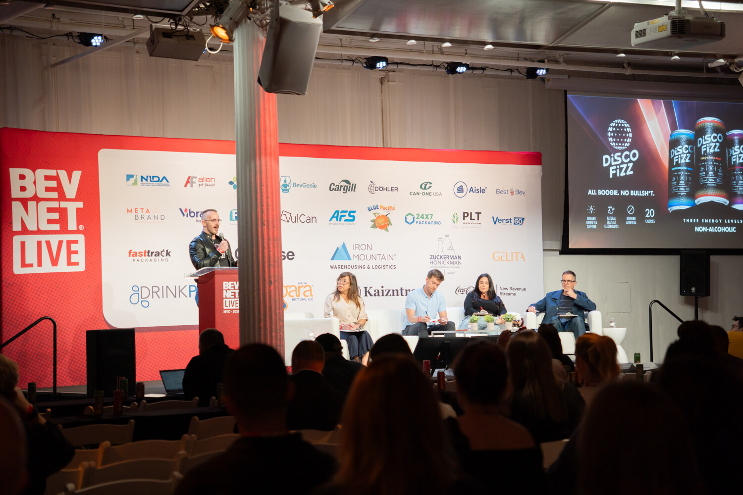 Wide view from the back of a dark audience toward a red BevNET Live stage with a presenter, a Disco Fizz slide and judges on couches.