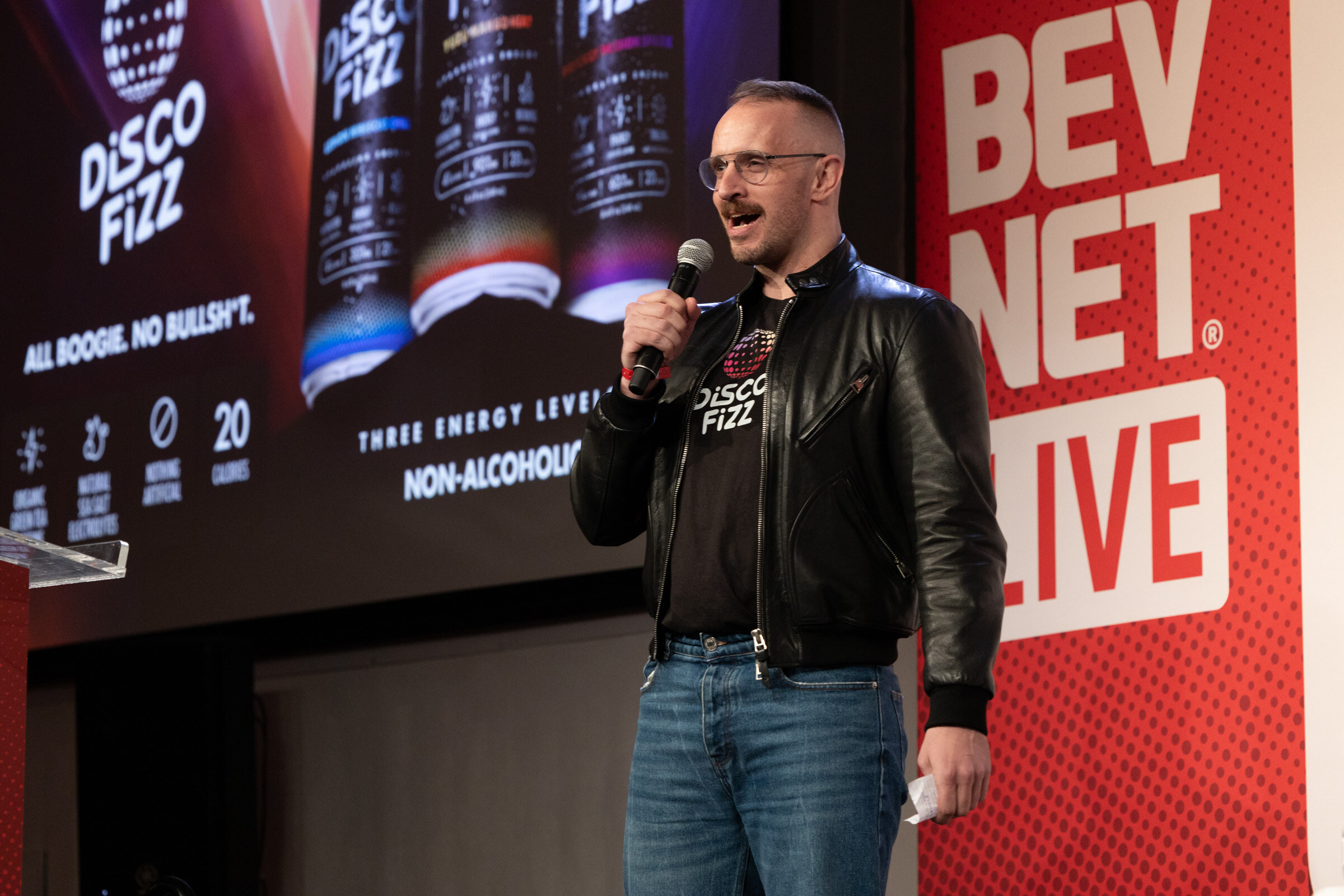 Man in a leather jacket and Disco Fizz T-shirt speaks into a microphone in front of a Disco Fizz slide and a BevNET Live banner.