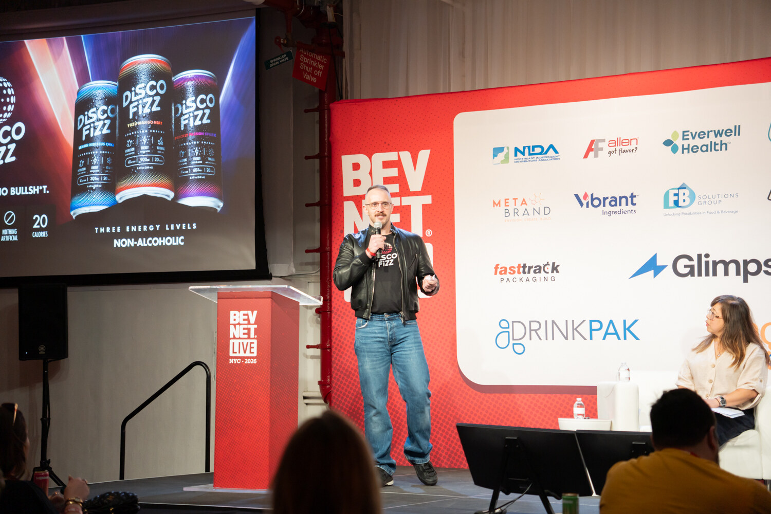 Presenter in a leather jacket on a red BevNET Live stage beside a podium, with Disco Fizz cans on the screen and sponsor logos.