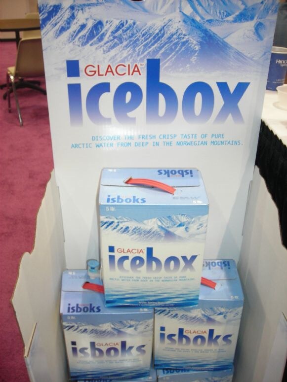 A cardboard display for Glacia Icebox shows a snowy mountain scene. Stacked 5-liter boxes of Icebox Arctic spring water with red carry handles sit in the display.