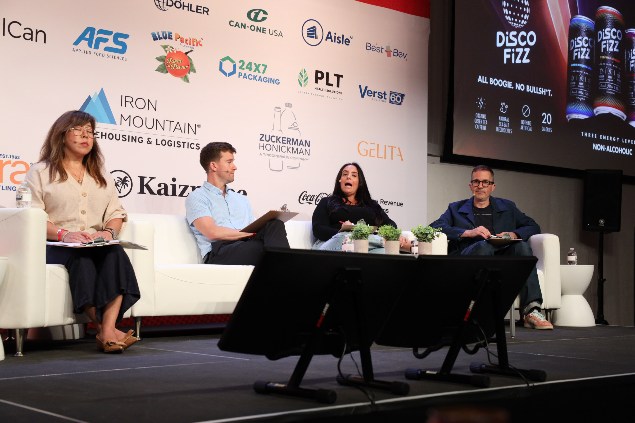 Four judges seated on white couches on a stage, with a sponsor wall of logos and a Disco Fizz slide on screen behind them.