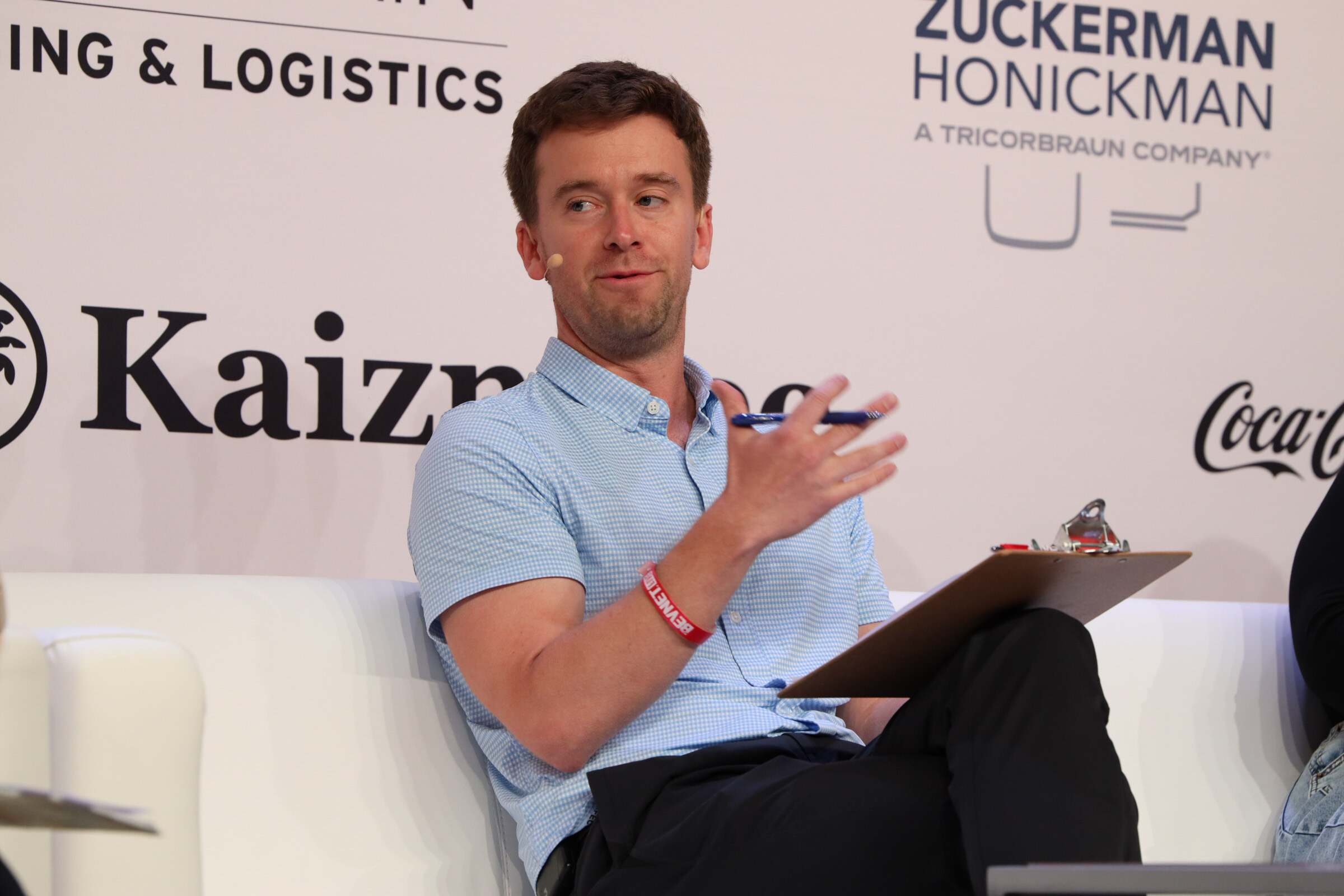 Man in a light blue short-sleeved shirt gestures while speaking from a couch, a clipboard on his knee and a Kaizntree logo behind him.
