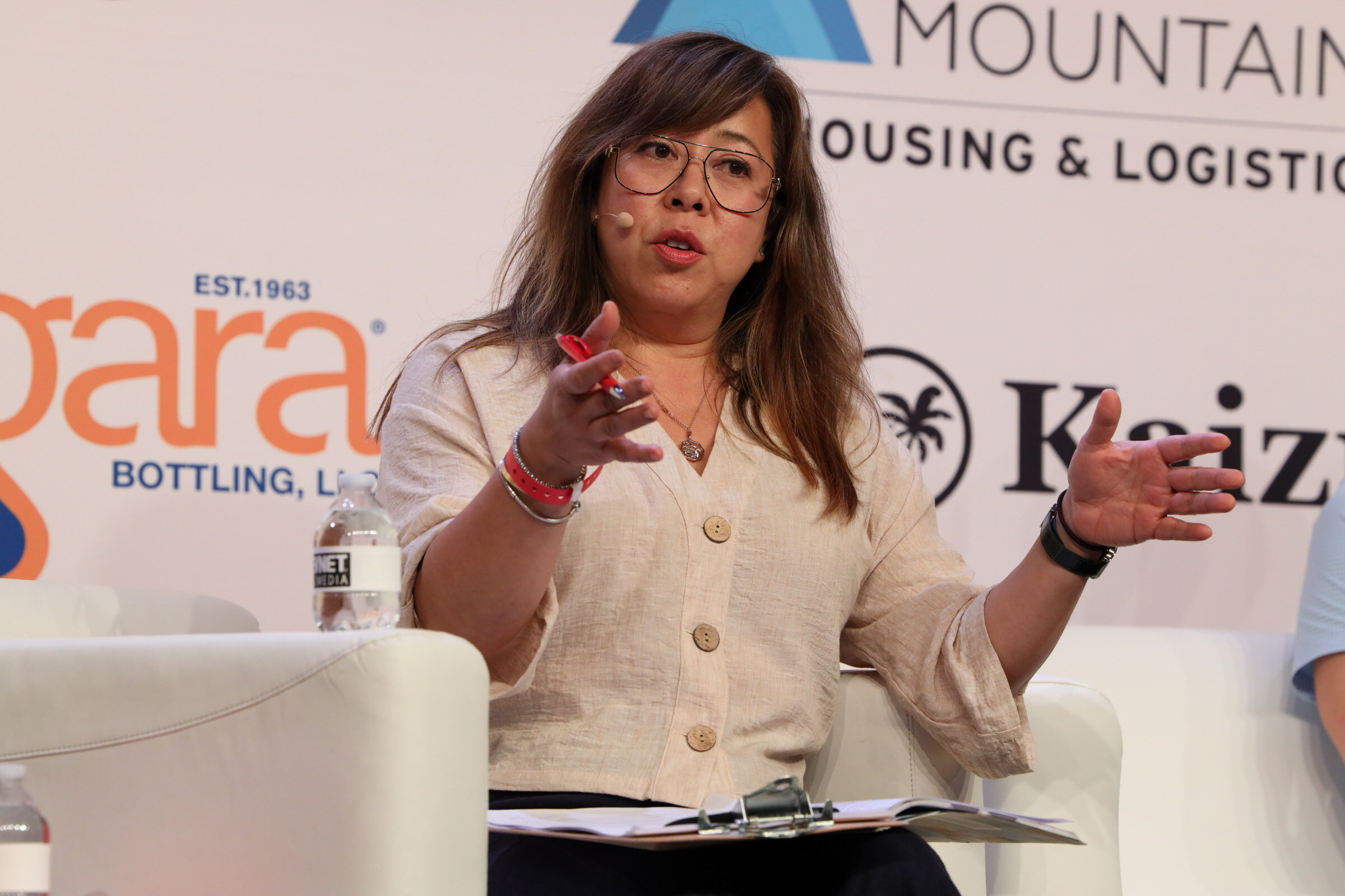Woman in glasses and a beige cardigan gestures with both hands while speaking from a couch in front of Niagara Bottling and Iron Mountain logos.