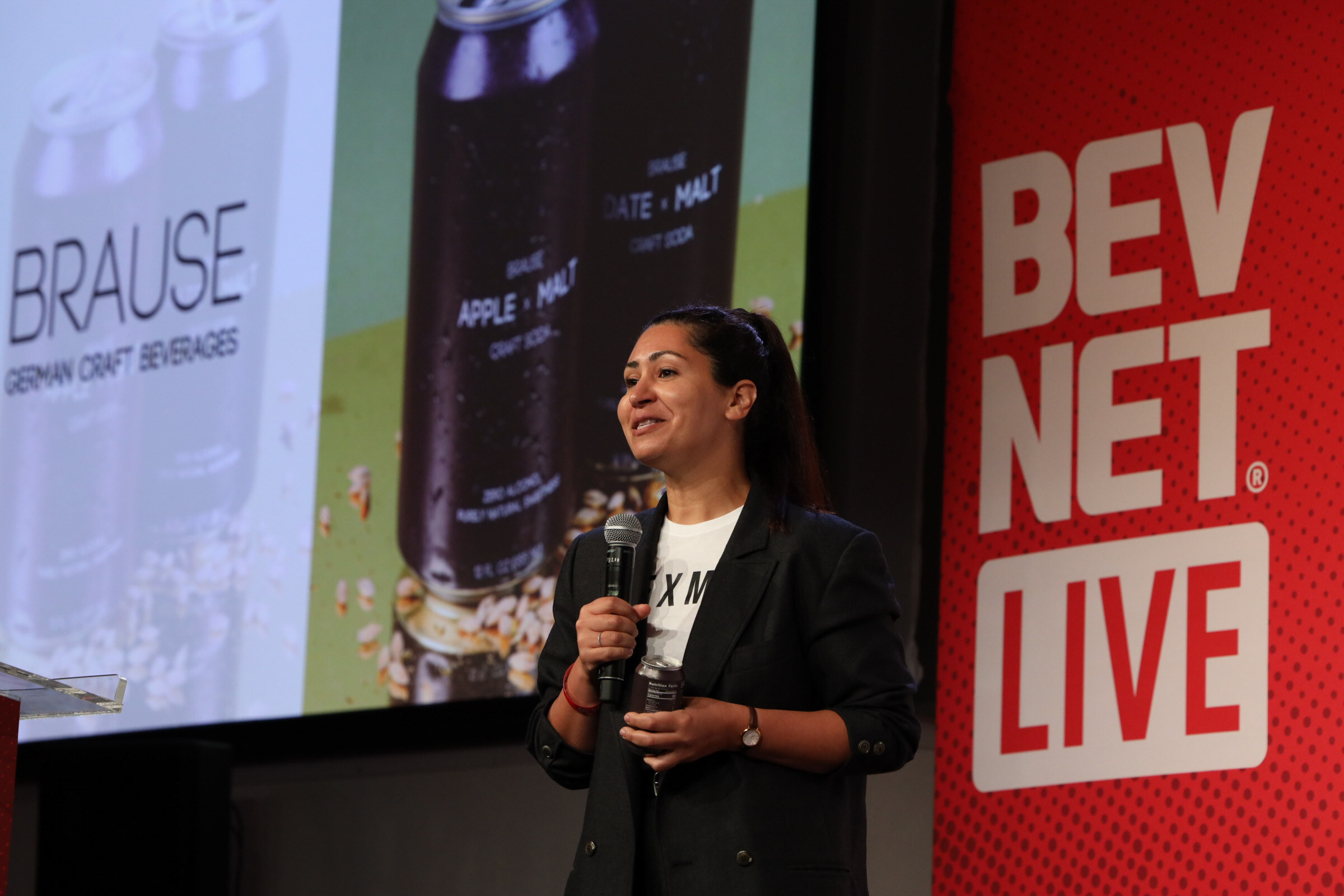 Woman in a black blazer speaks into a microphone holding a can, in front of a Brause German Craft Beverages slide.
