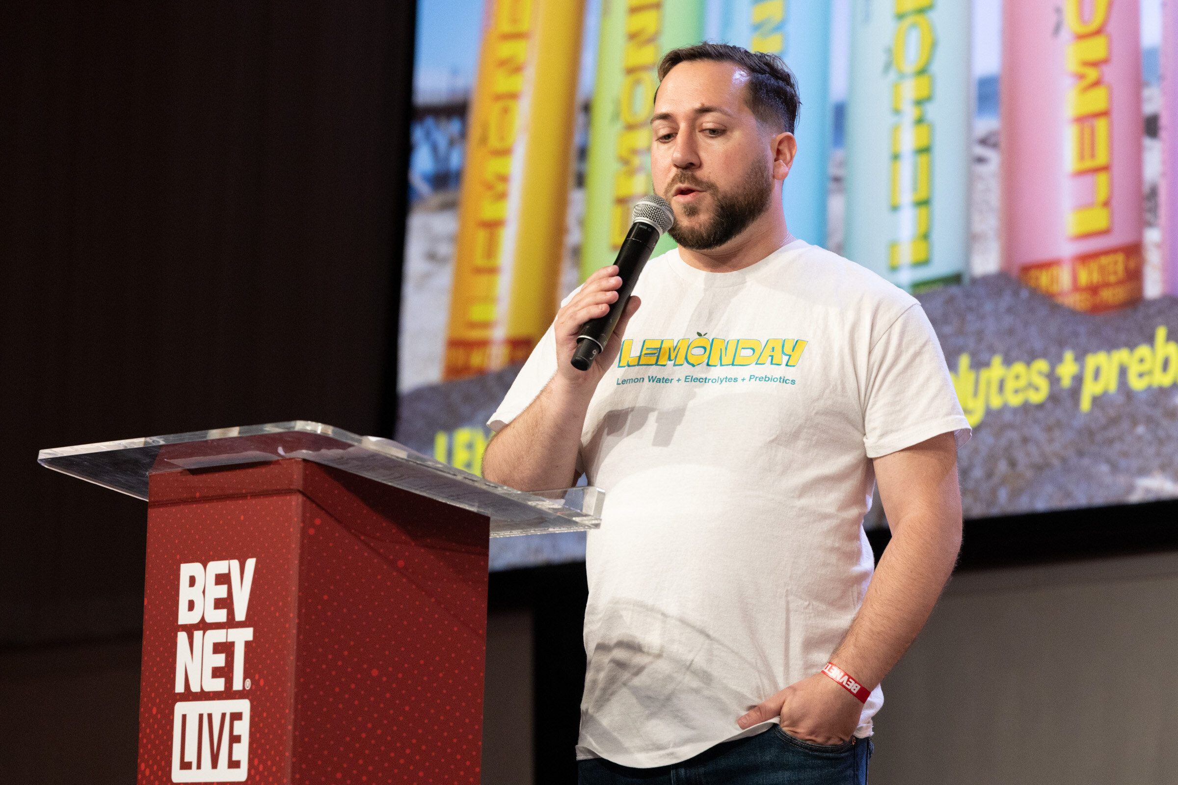 Man in a white Lemonday T-shirt speaks into a microphone, one hand in his pocket, beside the podium.