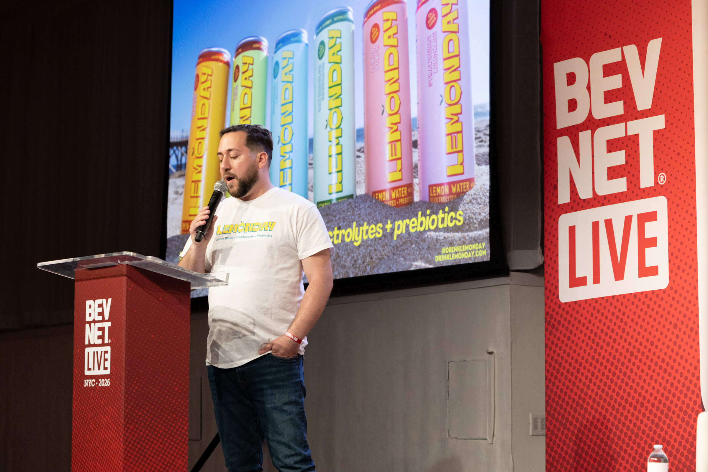 Man in a Lemonday T-shirt speaks at a podium in front of a slide of colorful Lemonday cans on a beach.