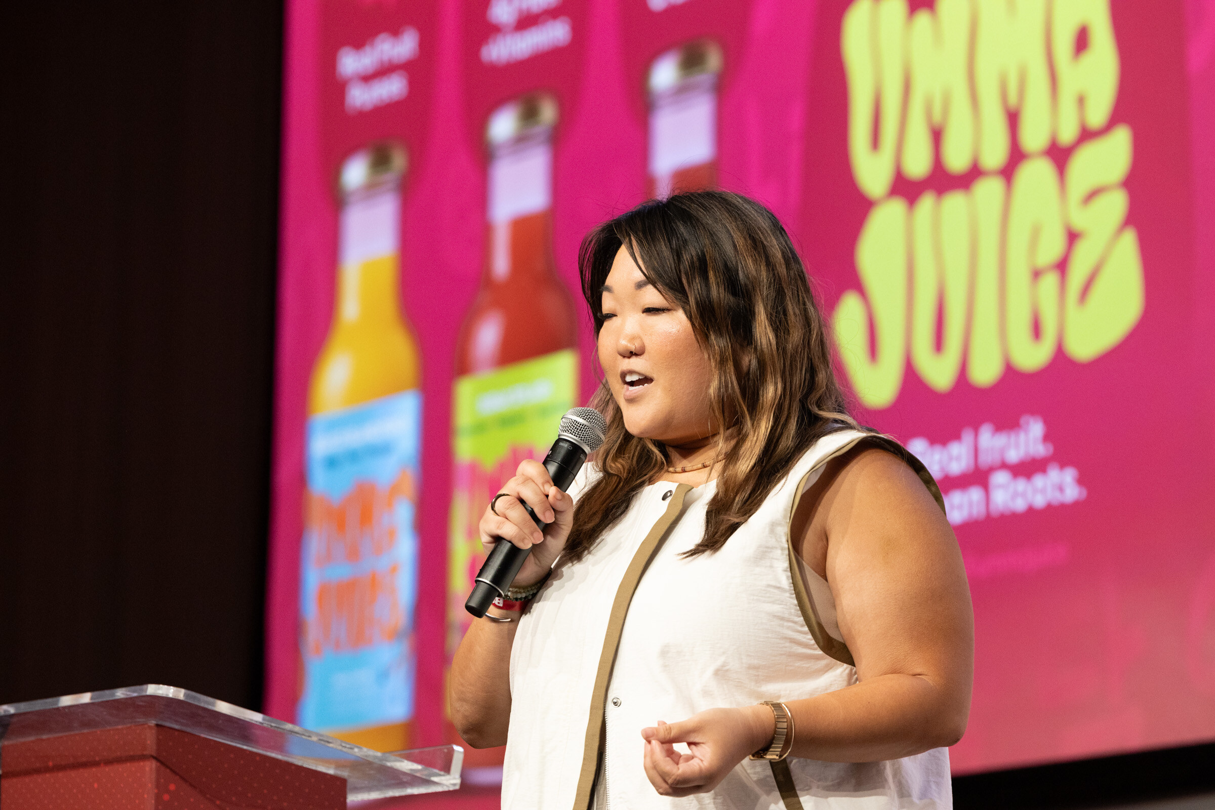 Woman in a white vest speaks into a microphone in front of a pink Umma Juice slide with three bottles.