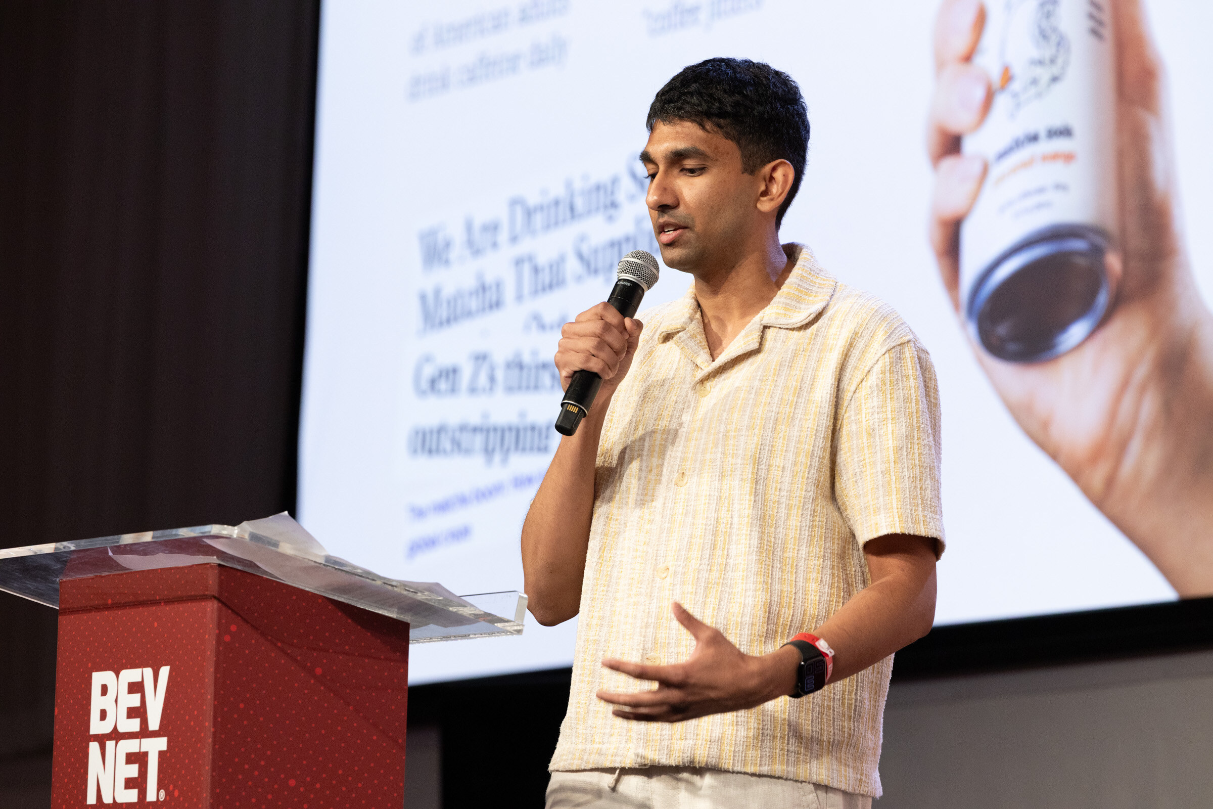 Man in a cream shirt gestures while speaking into a microphone in front of a slide about matcha demand.