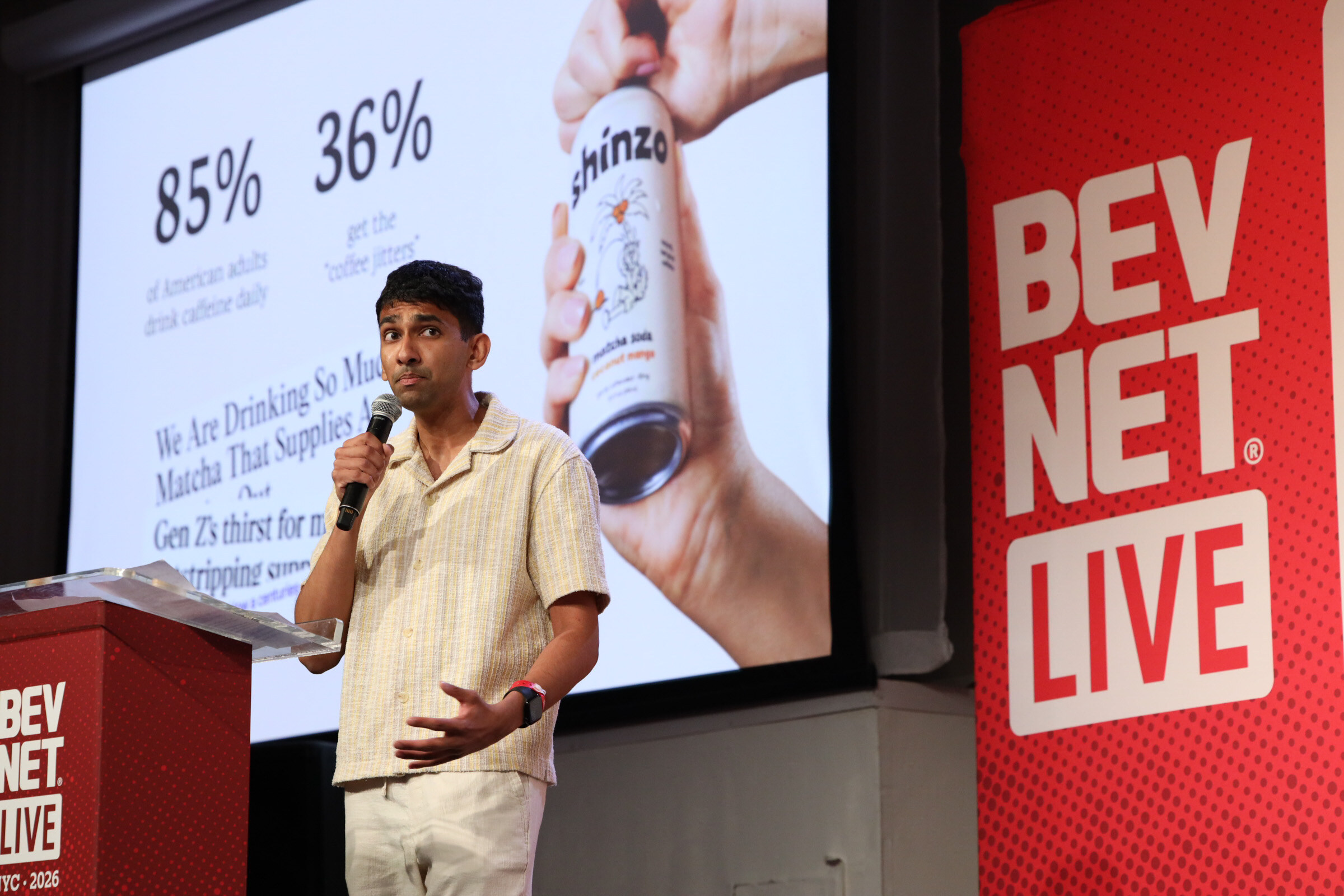 Man in a cream short-sleeved shirt speaks into a microphone beside a slide showing a hand holding a Shinzo matcha soda can.