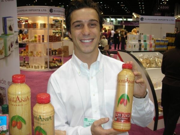 A smiling man in a white shirt holds a bottle of Gojilania goji juice, with more bottles in front of him and European Imports Ltd. shelves and signs behind him.