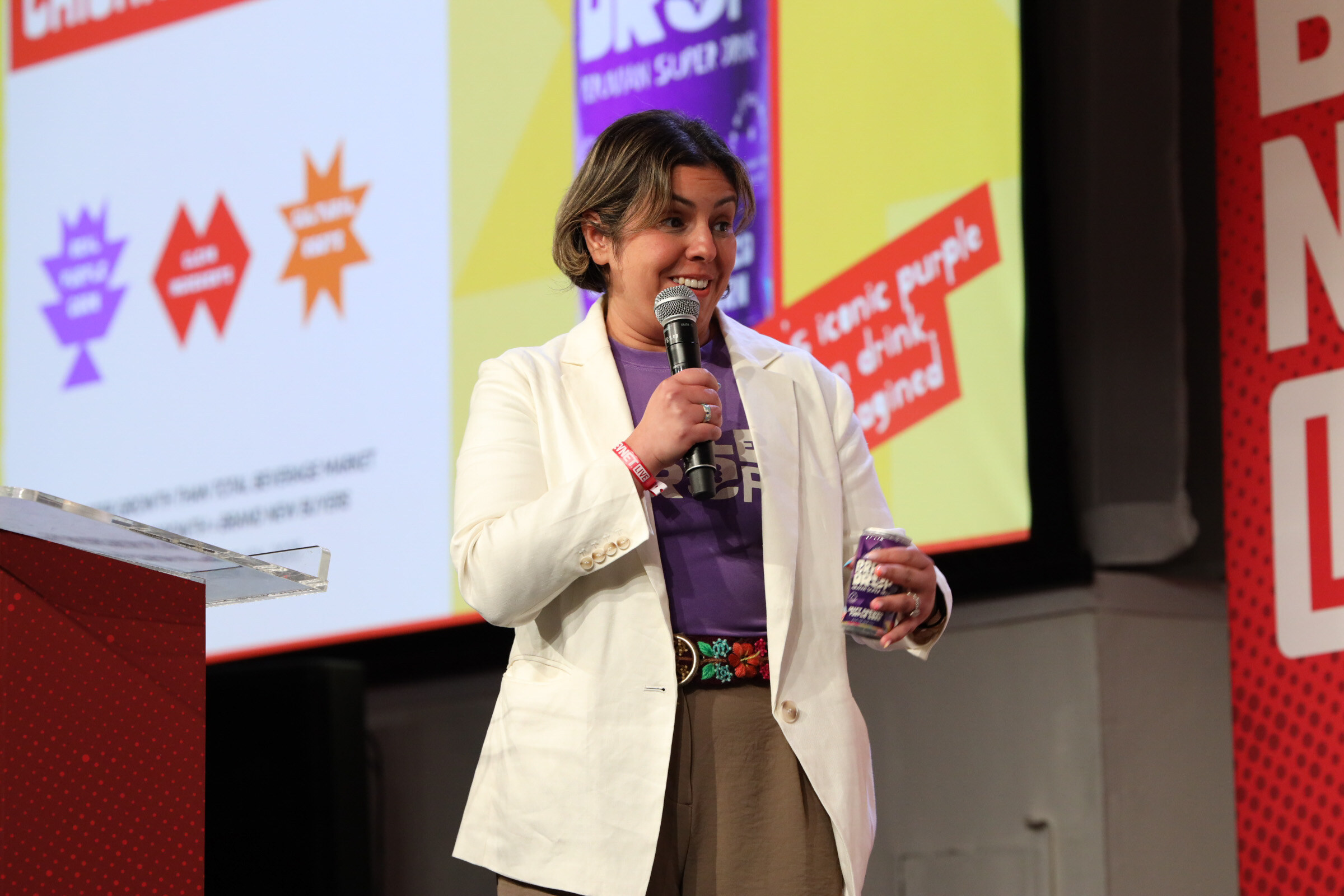 Smiling woman in a white blazer and purple Purple Drop T-shirt holds a can and a microphone in front of a yellow and purple slide.