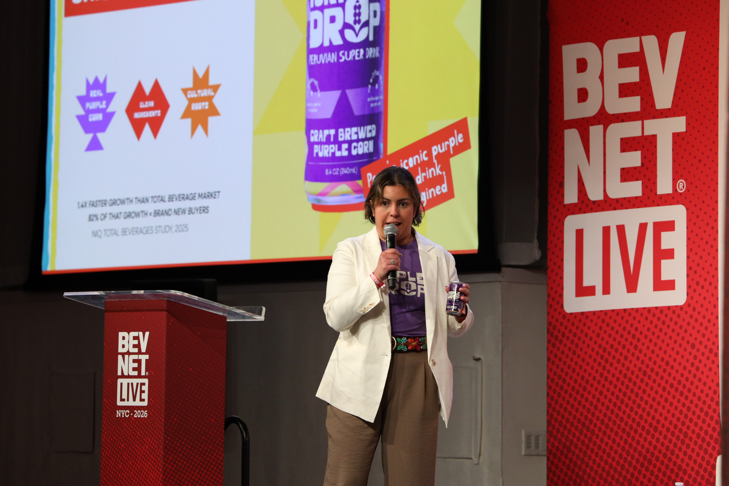 Presenter in a white blazer holding a purple can stands beside a BevNET Live podium under a slide of a Purple Drop can.