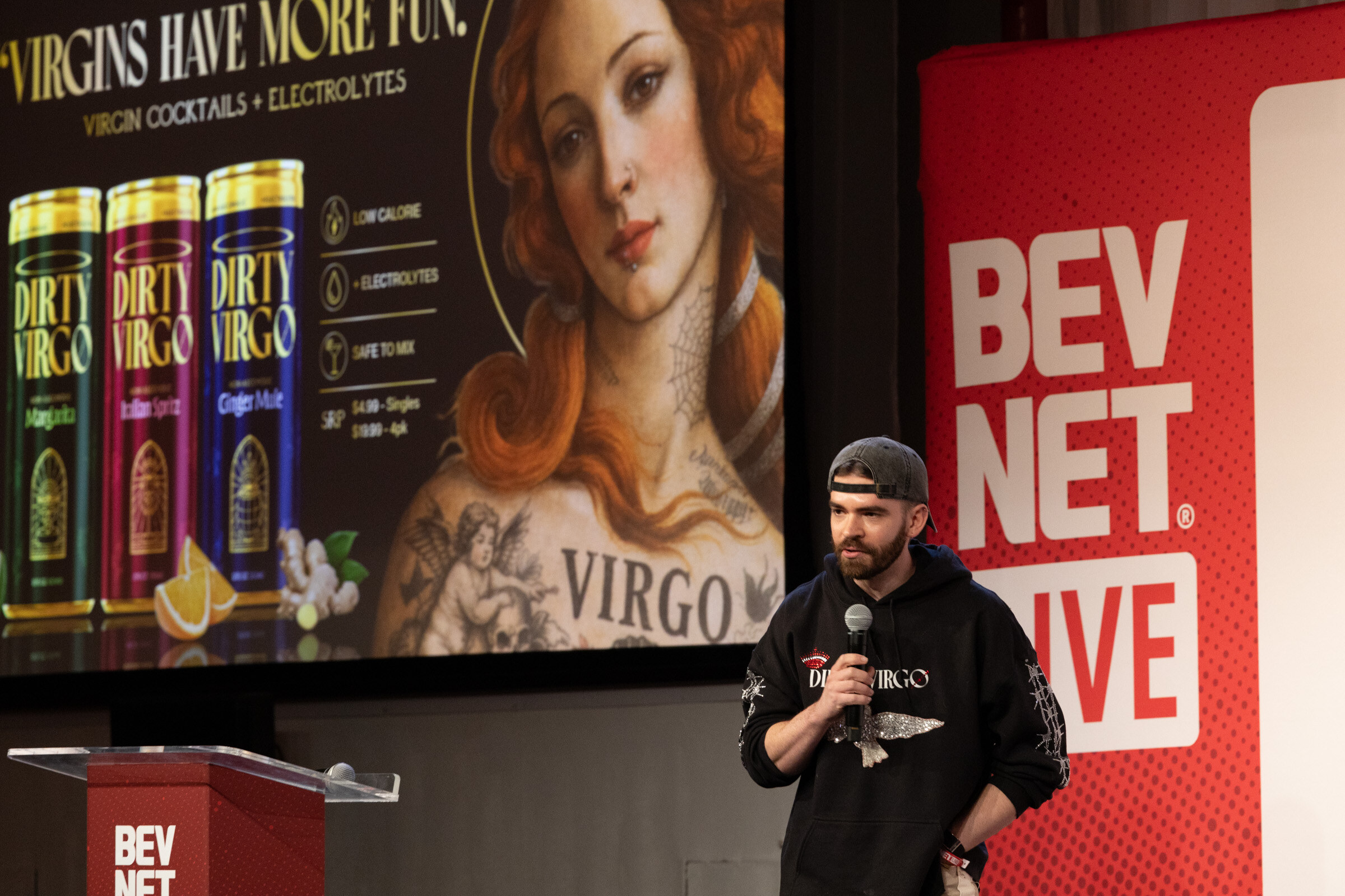 Man in a Dirty Virgo hoodie and backwards cap speaks beside a slide reading Virgins Have More Fun with three Dirty Virgo cans.