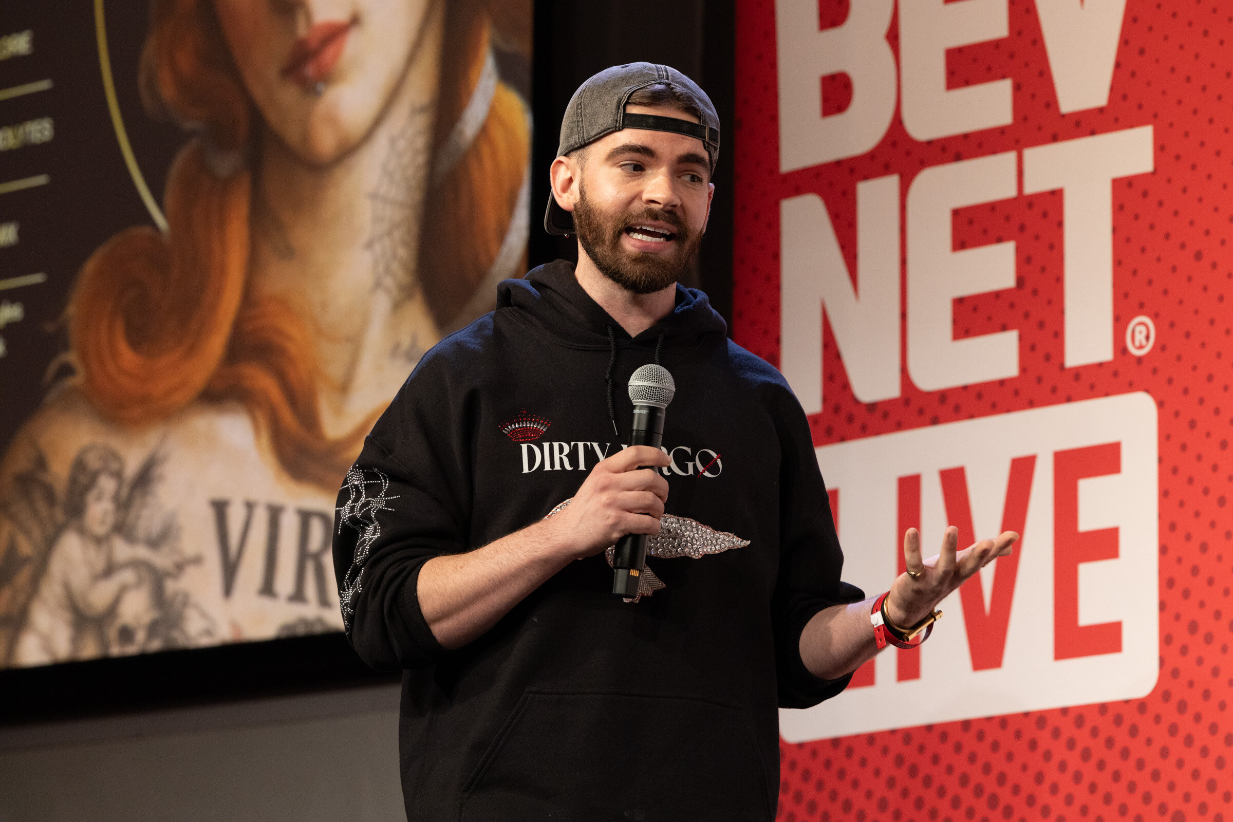 Man in a black Dirty Virgo hoodie gestures while speaking into a microphone in front of a BevNET Live banner.