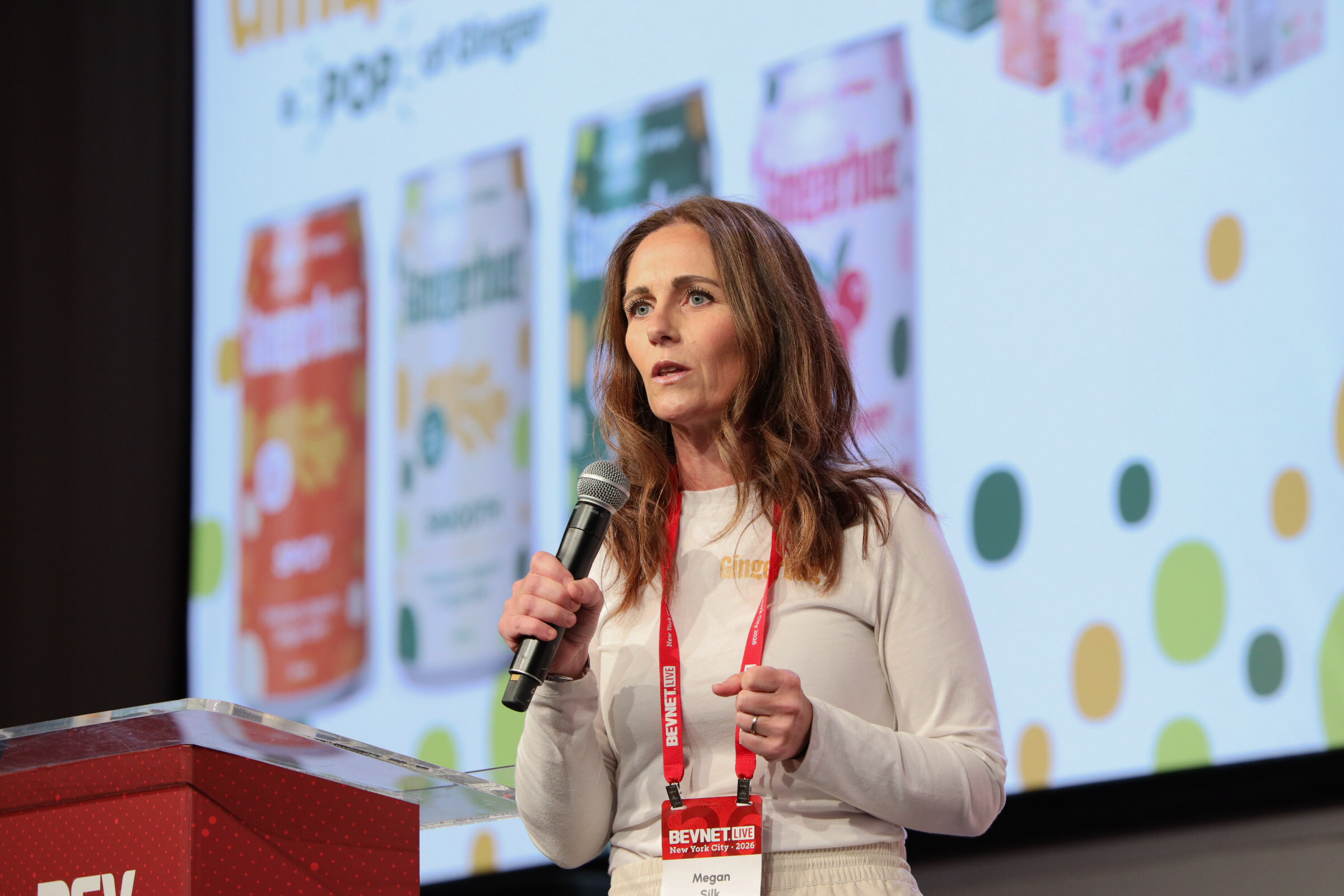 Woman with a BevNET Live lanyard speaks into a microphone in front of a blurred slide of Gingerbug cans.