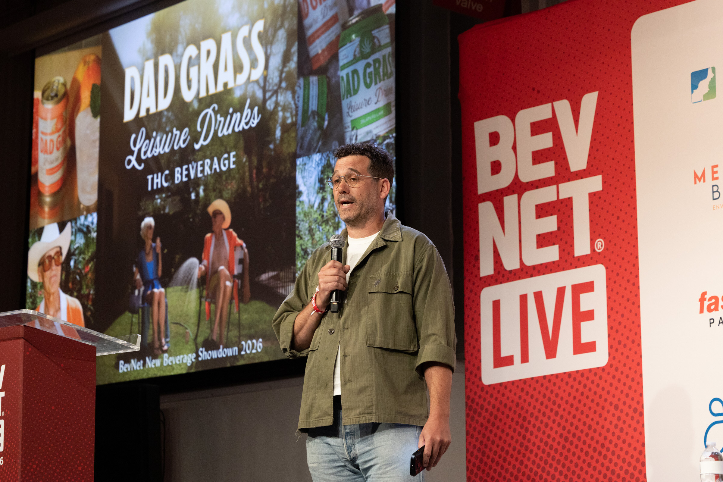 Man in an olive overshirt speaks into a microphone in front of a Dad Grass Leisure Drinks THC Beverage slide.
