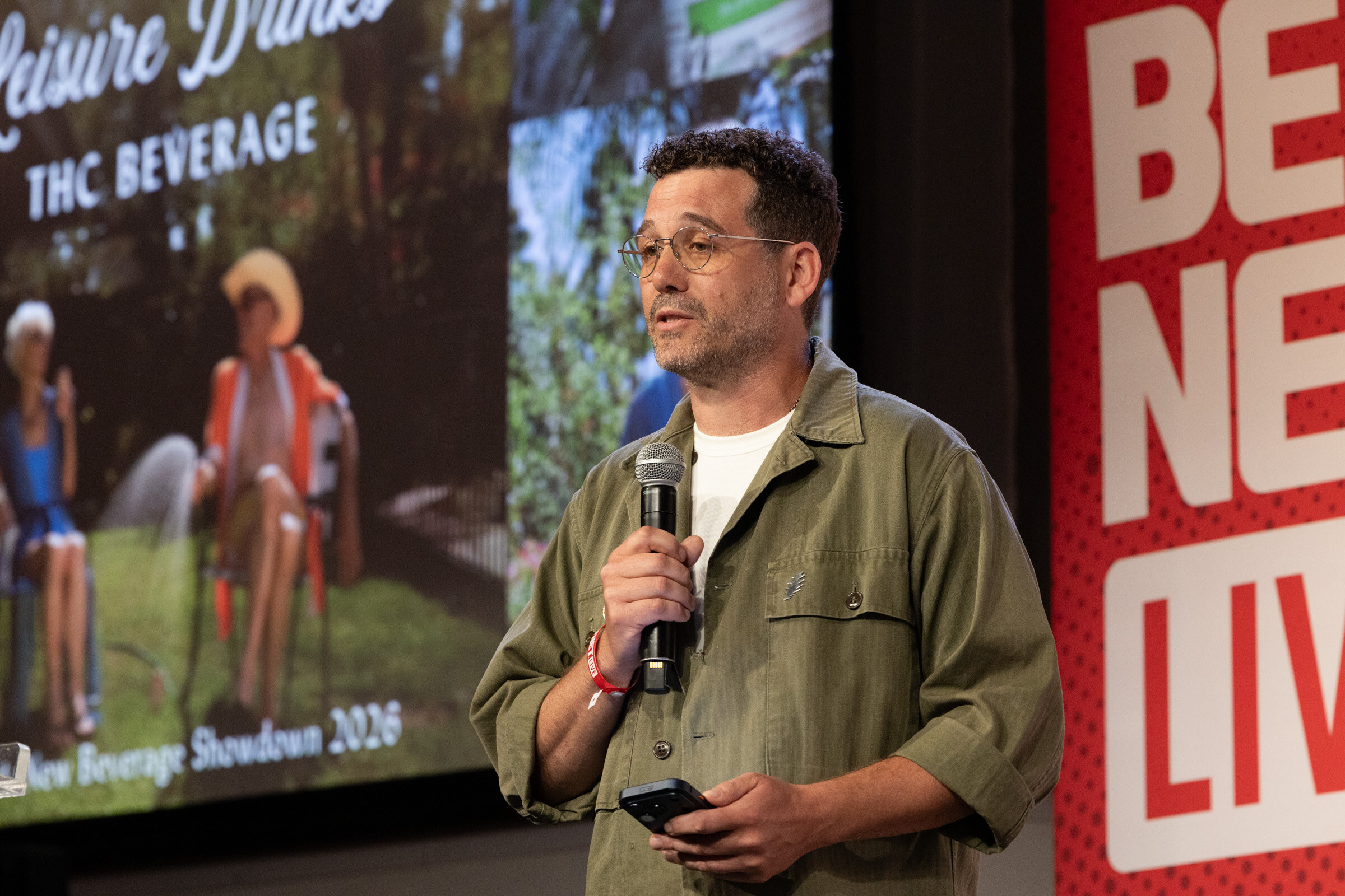 Man in glasses and an olive overshirt speaks into a microphone, holding a clicker, in front of a garden-party slide.