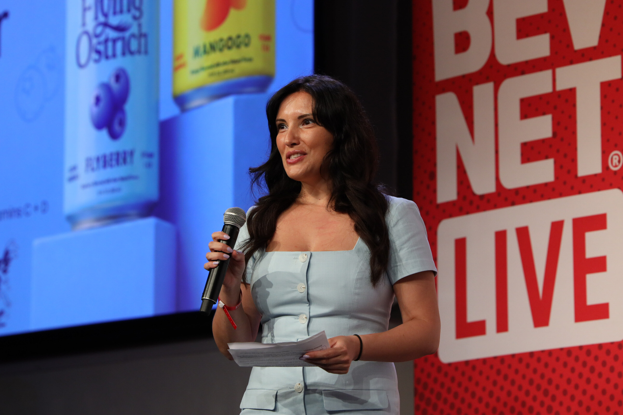 Smiling woman in a light blue dress holds a microphone and notes in front of a slide of Flying Ostrich Flyberry and Mangogo cans.