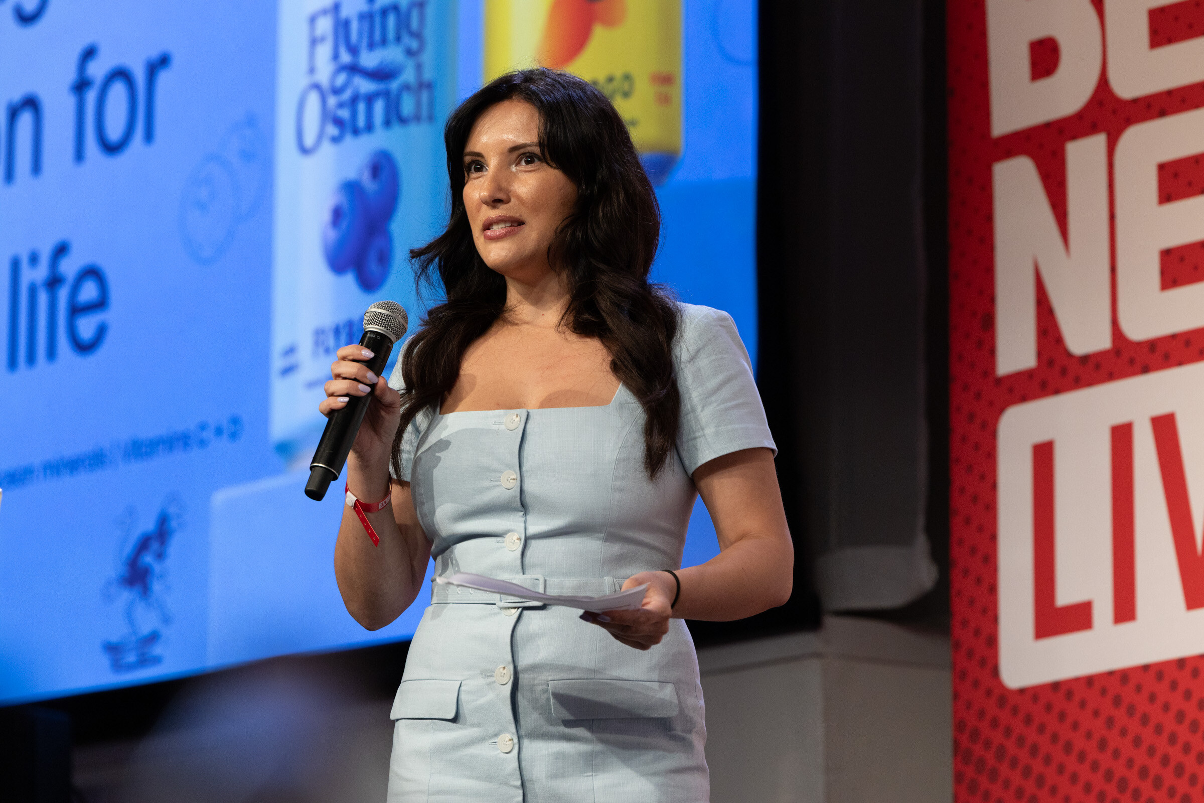 Woman in a light blue dress speaks into a microphone in front of a blue Flying Ostrich slide and a BevNET Live banner.