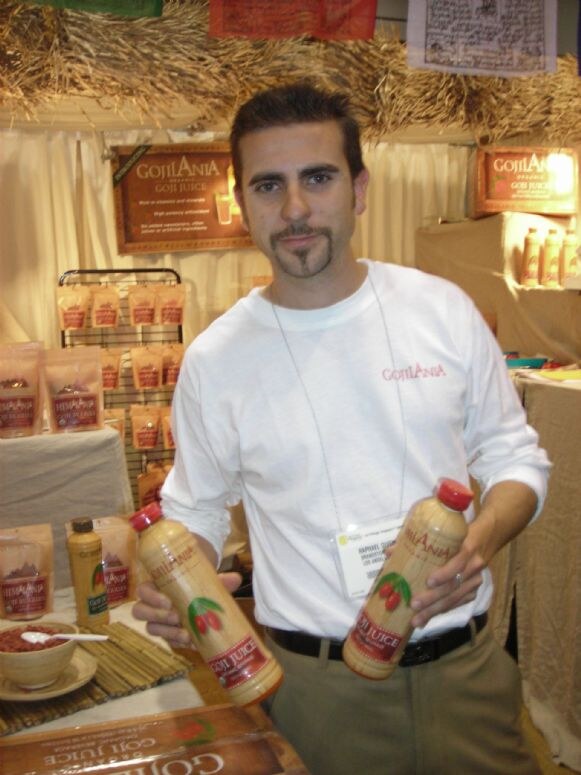 A man with a goatee and a white Gojilania shirt holds two bottles of GojiLania goji juice in a booth decorated with straw, signs and bags of dried goji berries.