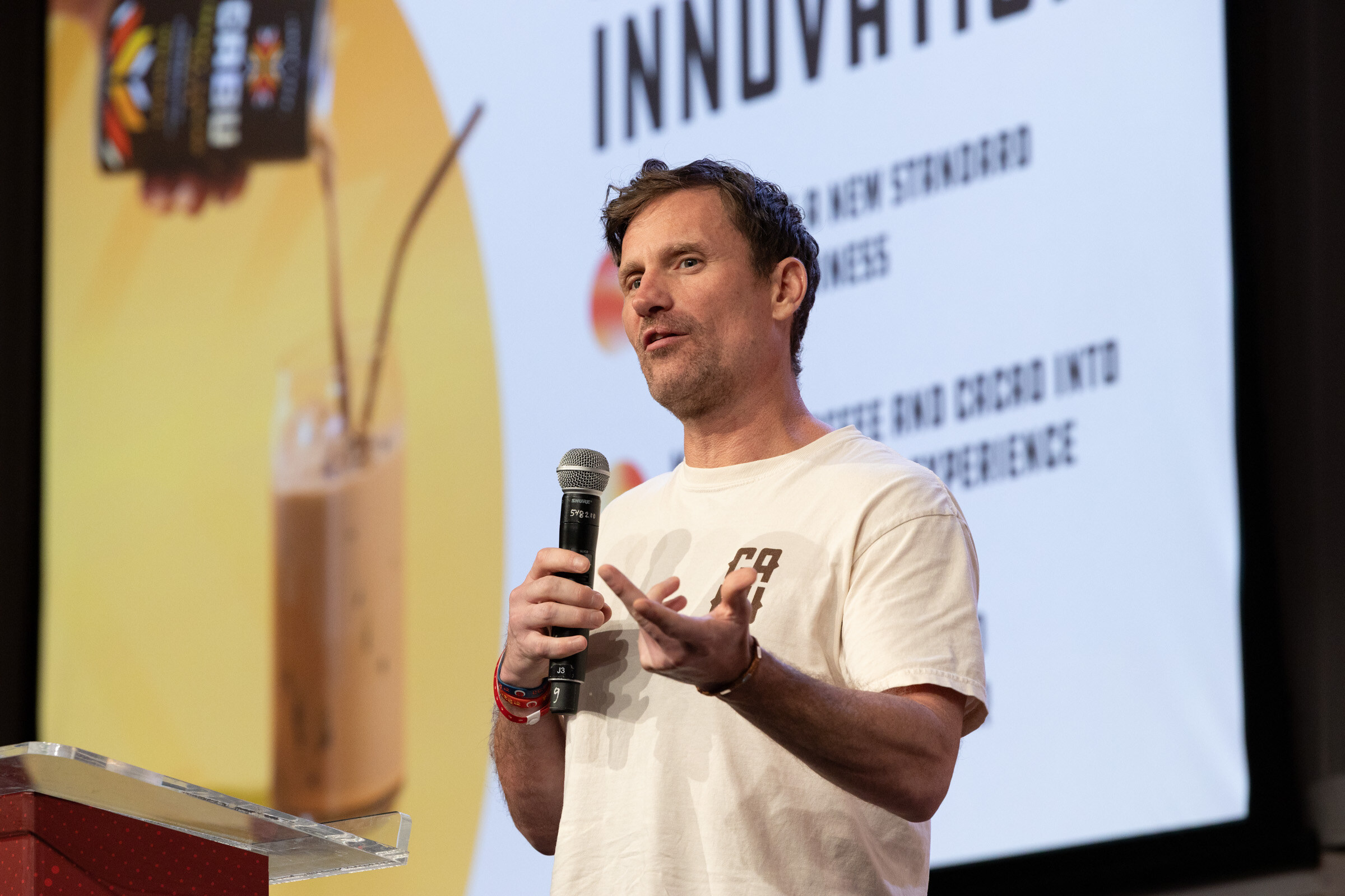 Man in a cream Cabu T-shirt gestures while speaking into a microphone in front of a slide of an iced coffee drink.