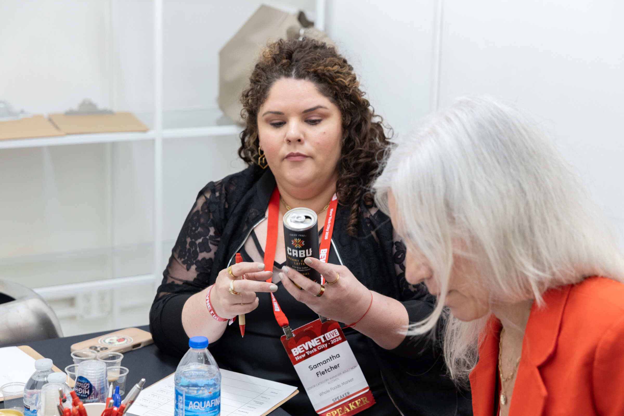 Woman with curly hair and a BevNET Live speaker lanyard holds a can of Cabu at a judging table beside another judge.
