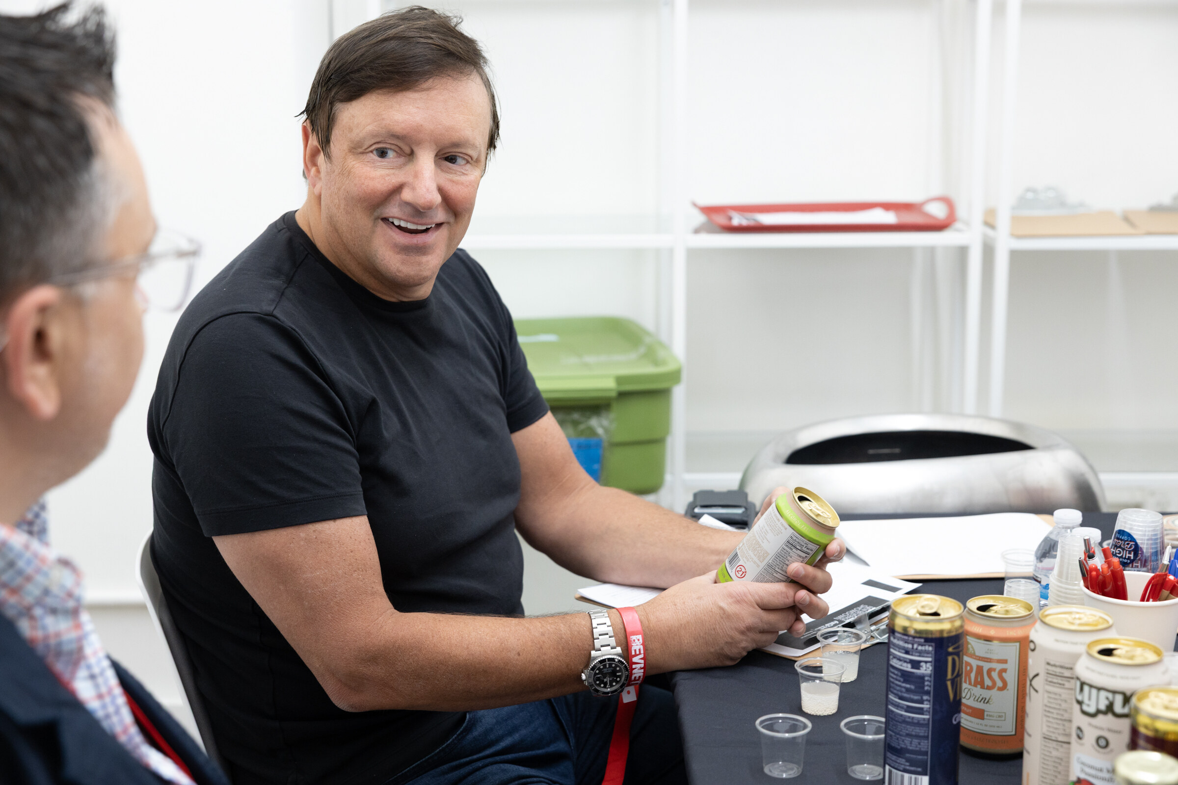 Man in a black T-shirt smiles while holding a can at a table crowded with cans, including Dad Grass and Lyflo.