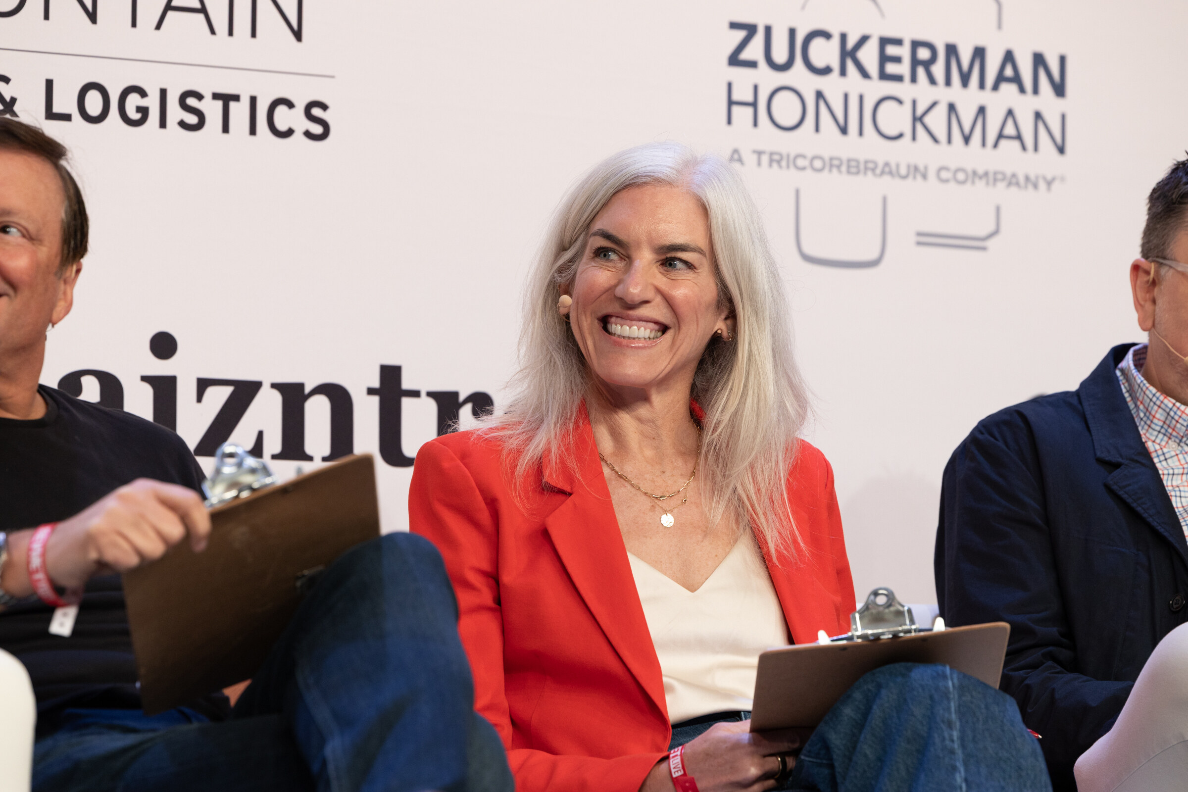Smiling woman with long silver hair in a red blazer sits on a couch holding a clipboard.