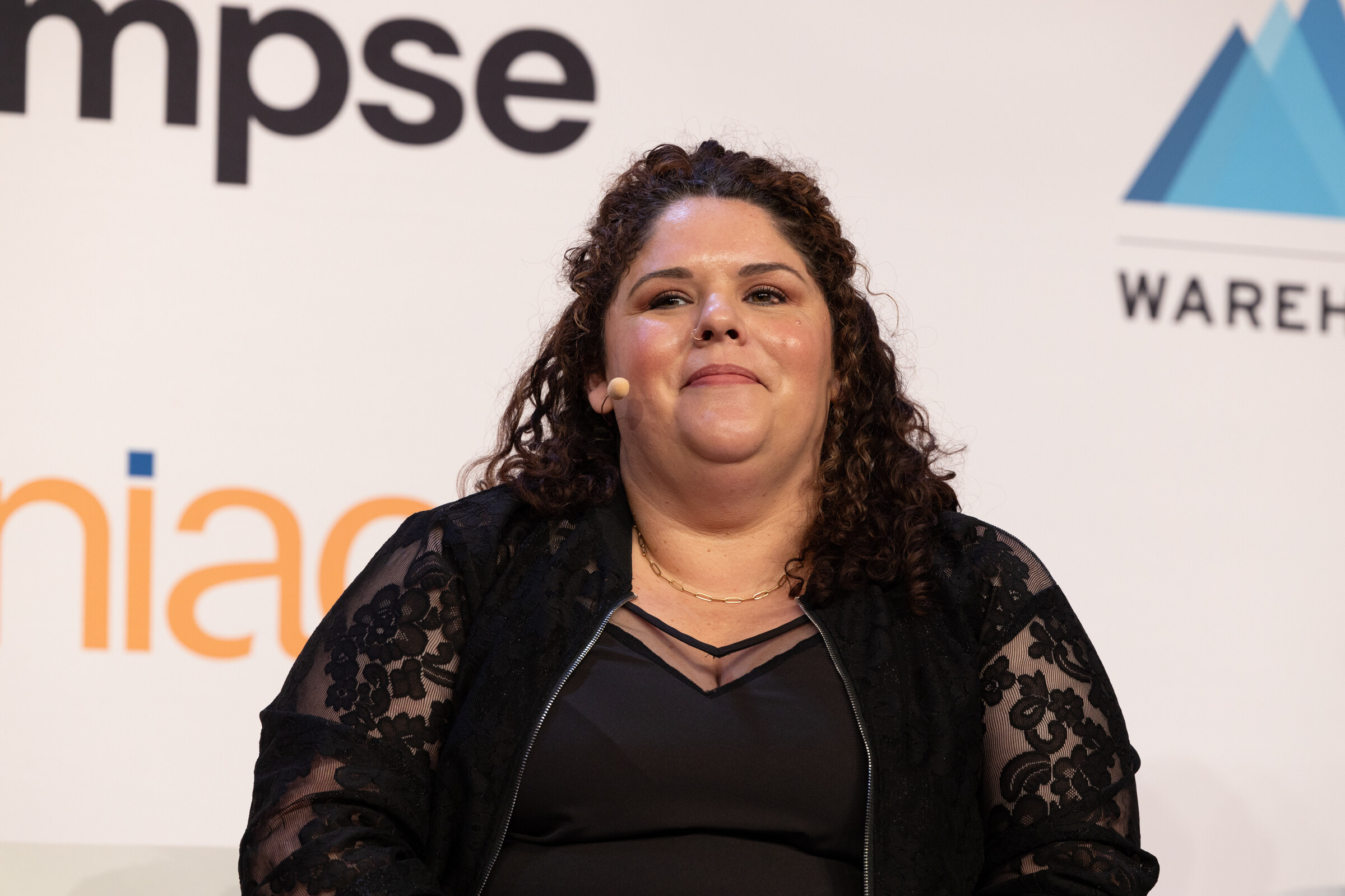 Woman with curly dark hair in a black lace top sits on stage in front of Glimpse and Niagara logos.