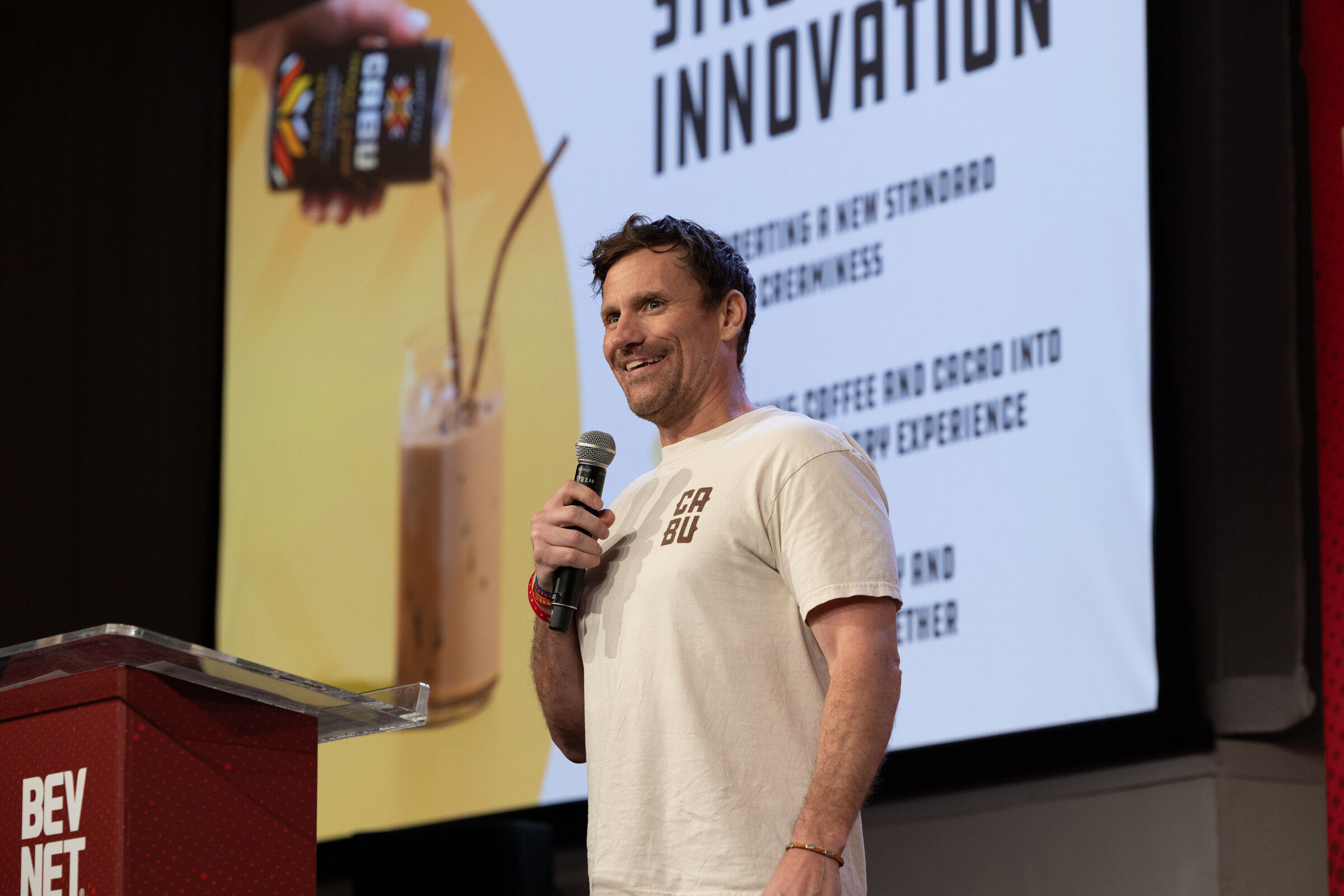 Smiling man in a Cabu T-shirt holds a microphone beside a Structural Innovation slide showing a Cabu carton and an iced drink.